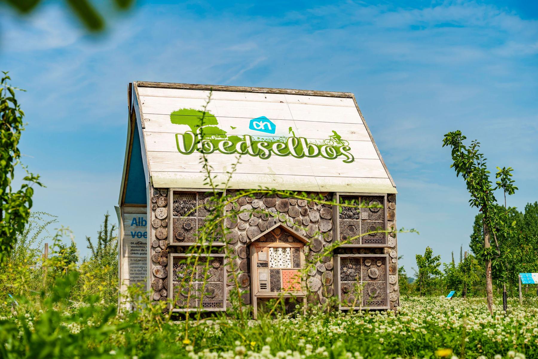 Kleines Haus mit Holzstämmen und Insektenhotels an der Fassade, Schriftzug 'voedselbos' auf dem Dach, umgeben von grüner Wiese und Bäumen