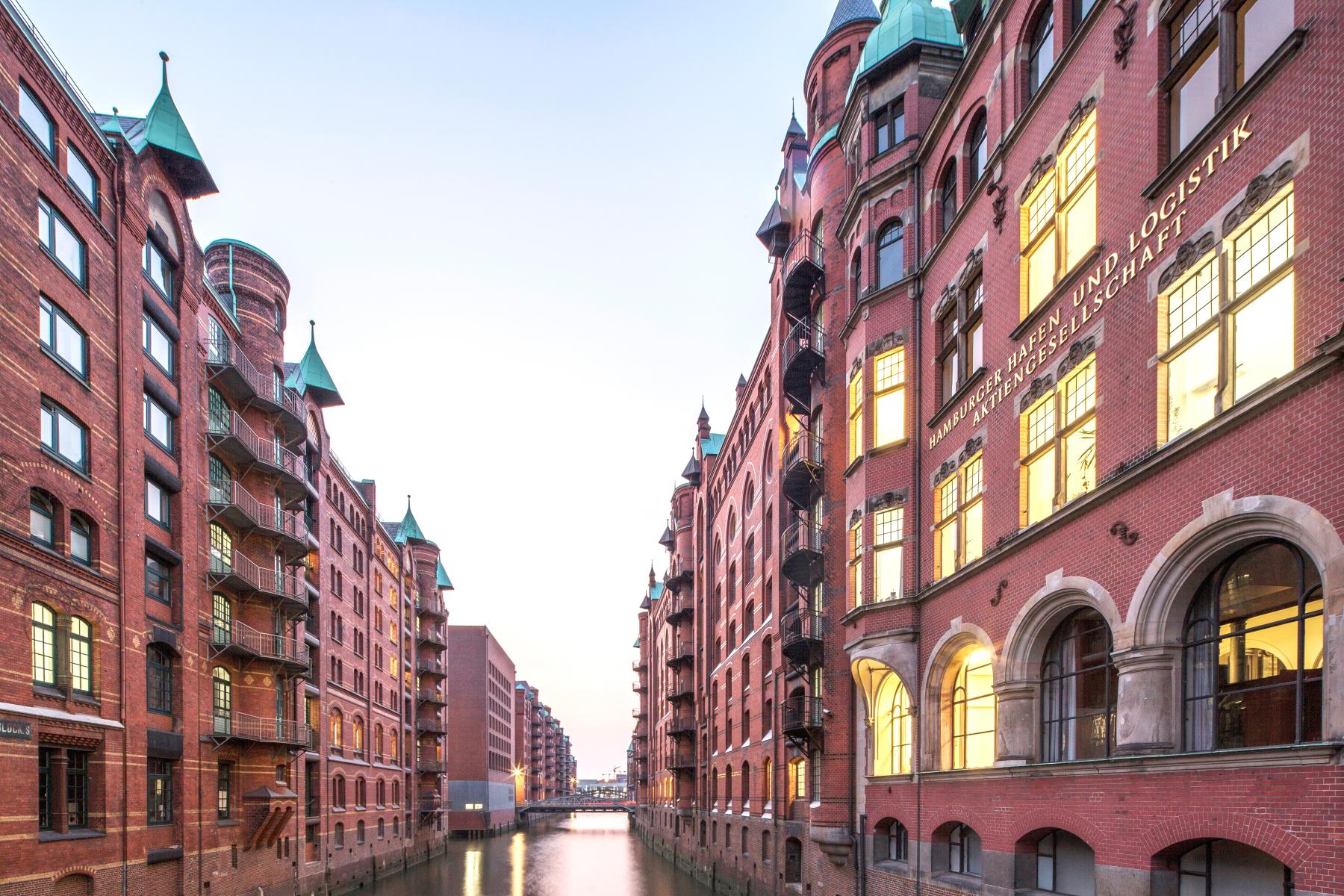 Zwei Backsteingebäude mit grünen Türmchen an einem Kanal in der Speicherstadt, Hamburg. Sichtbare Fenster und reflektierendes Wasser.