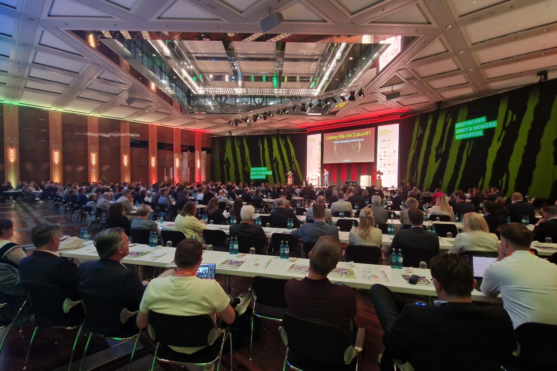 Großer Konferenzsaal mit vielen sitzenden Teilnehmern, Bühne mit Präsentationsbildschirm, grün beleuchtete Wände mit dem Logo "Deutscher Obst und Gemüse Kongress".