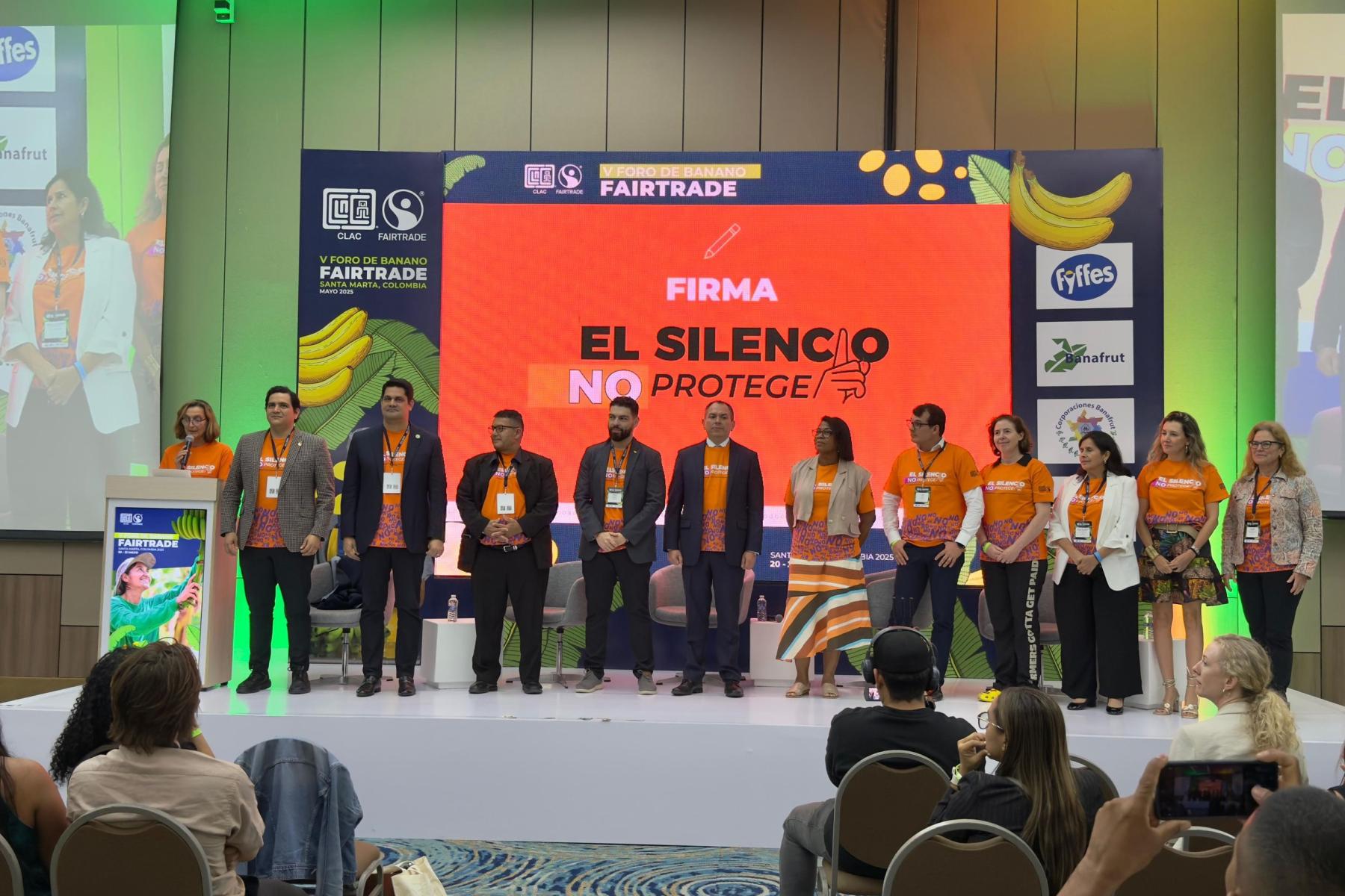 Eine Gruppe von Personen auf einer Bühne, alle tragen orangefarbene T-Shirts mit dem Text 'El Silencio No Protege'. Im Hintergrund sind Fairtrade-Logos und Bananenmuster sichtbar.