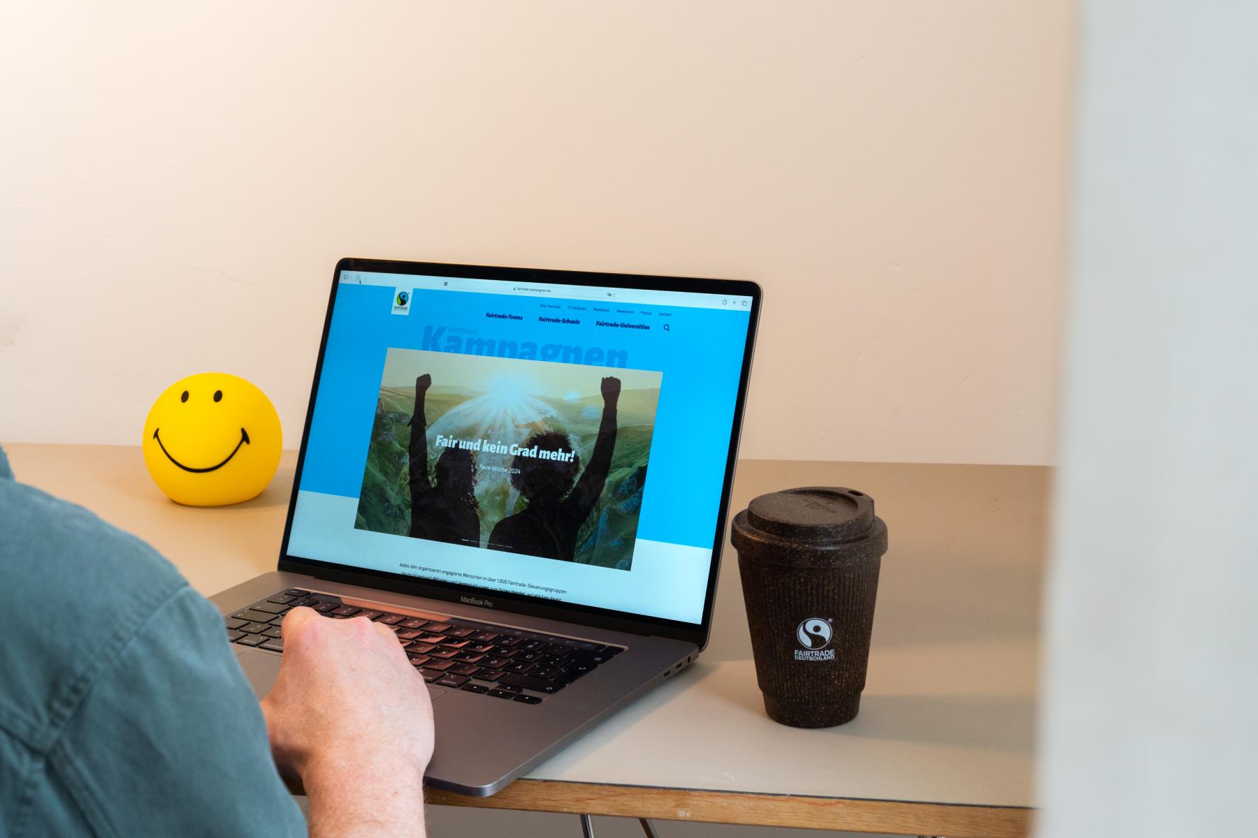 Eine Person sitzt an einem Laptop. Auf dem Bildschirm ist die Fairtrade Kampagnen Website zu sehen Neben dem Laptop steht ein Kaffeebecher mit Fairtrade-Logo und ein gelber Smiley