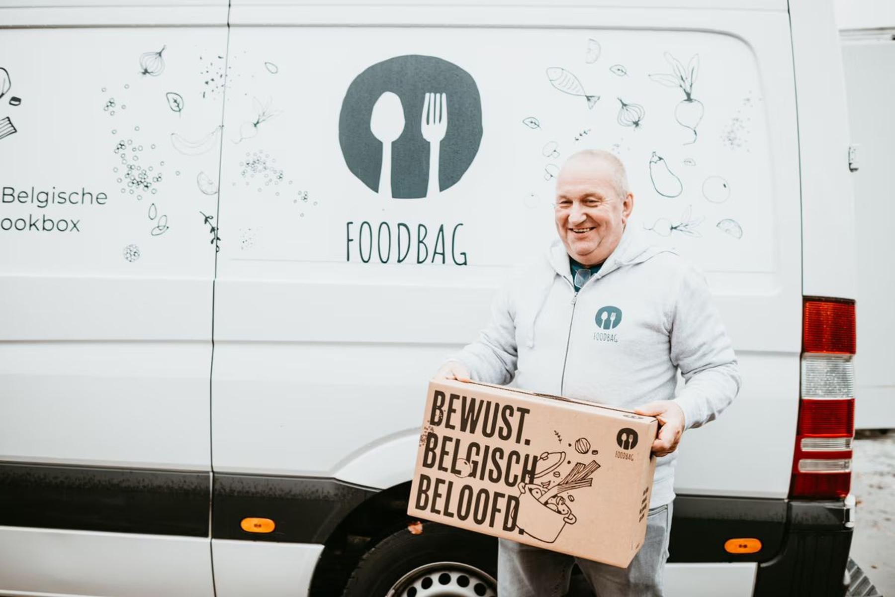 Eine Person hält eine große, braune Box mit dem Text 'BEWUST. BELGISCH. BELOOFD.' vor einem weißen Lieferwagen mit einem grafischen Design von Lebensmitteln und dem Logo 'FOODBAG'.