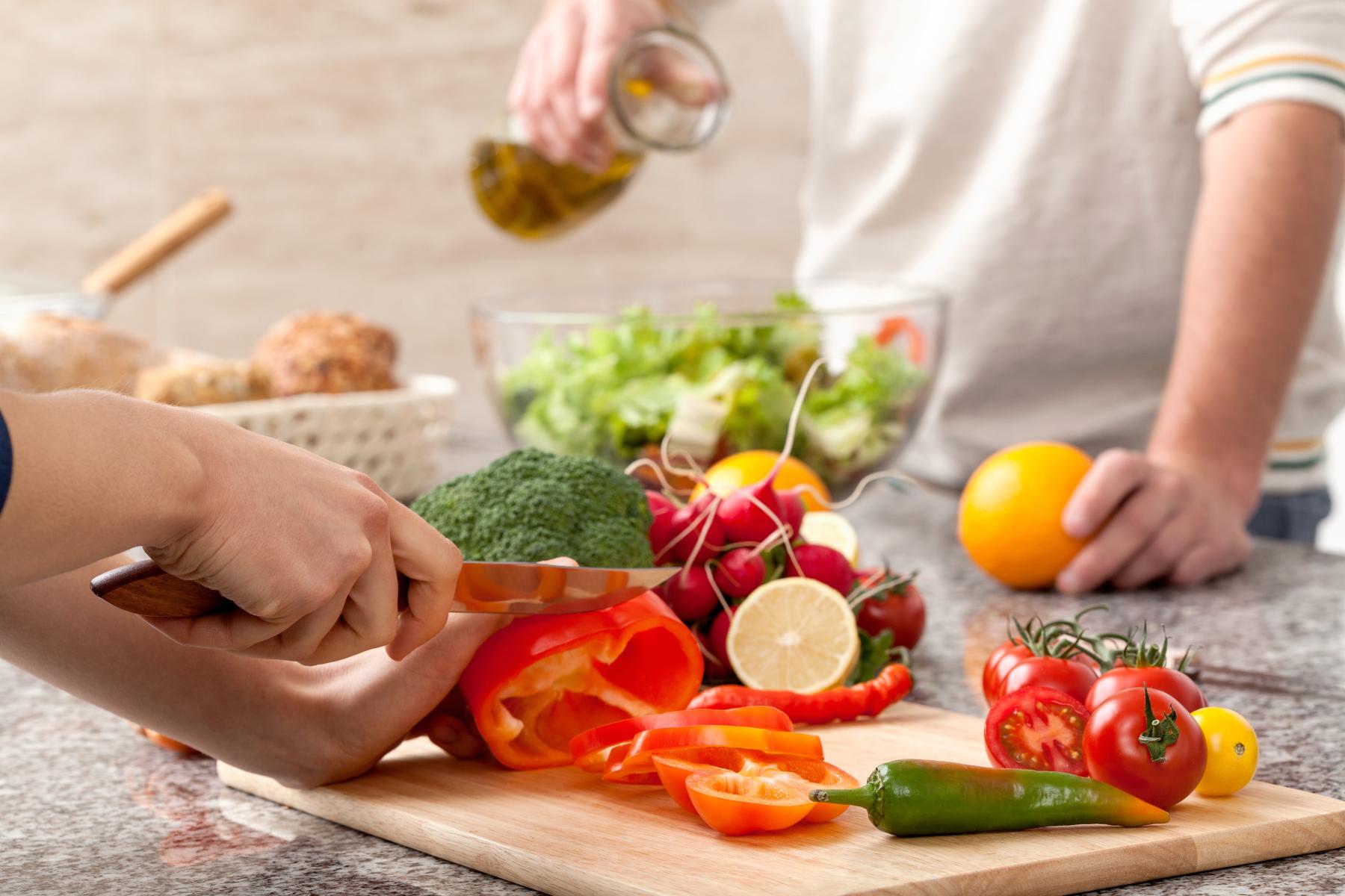 Eine Hand schneidet eine rote Paprika auf einem Holzschneidebrett. Im Hintergrund wird ein Salat in einer Schüssel zubereitet. Weitere Gemüse wie Brokkoli, Tomaten, eine Limette und eine Orange sind sichtbar.
