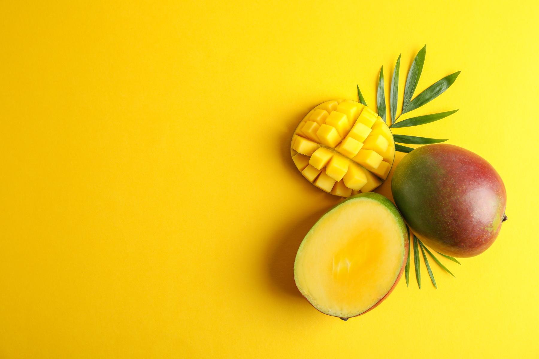 Eine halbierte Mango mit gelbem Fruchtfleisch und eine ganze Mango liegen auf einem gelben Hintergrund, umgeben von grünen Blättern.