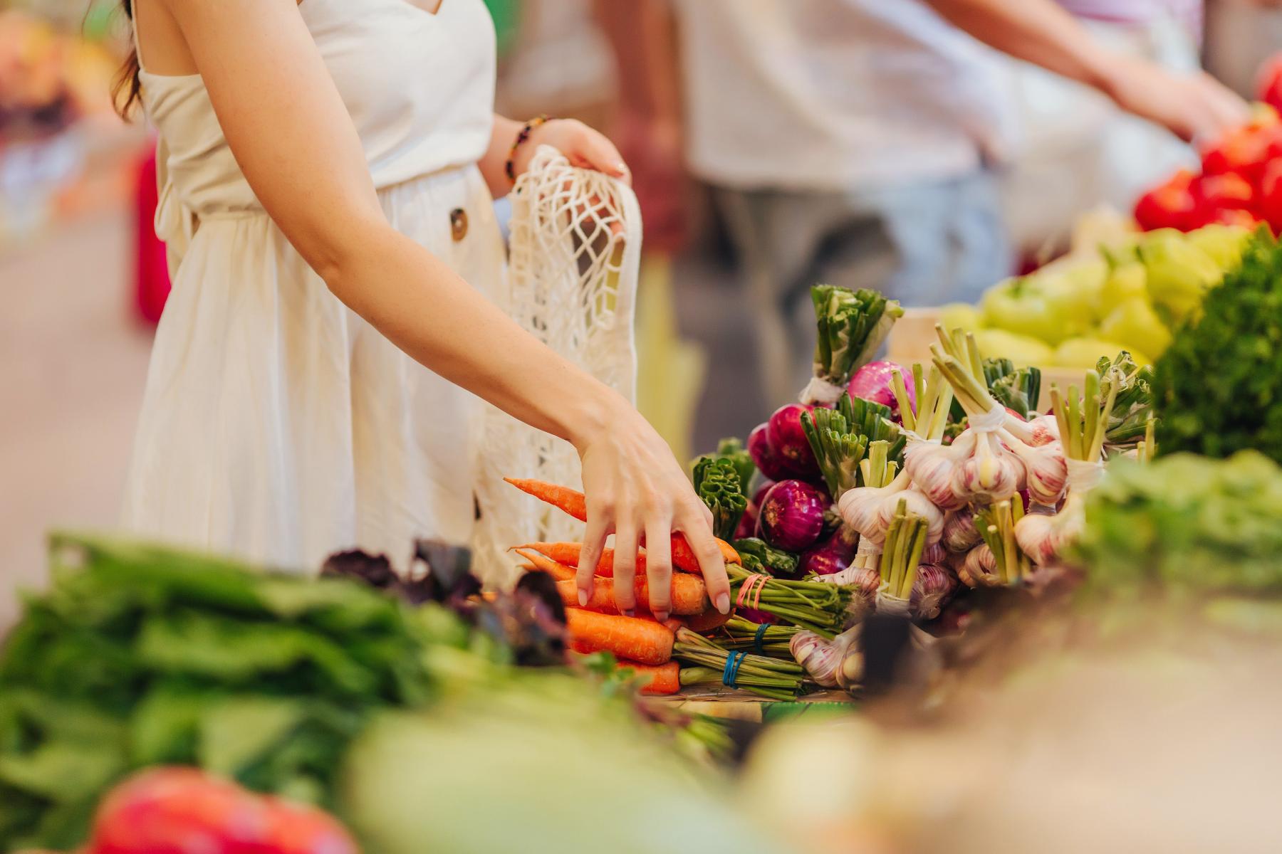 Eine Person in einem weißen Kleid greift nach Karotten auf einem Marktstand, umgeben von frischem Gemüse wie Knoblauch und Blattgemüse.