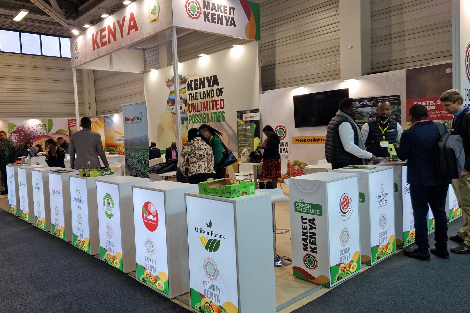 Messestand mit dem Titel 'MAKE IT KENYA'. Tische mit Logos von verschiedenen Unternehmen. Menschen interagieren am Stand.