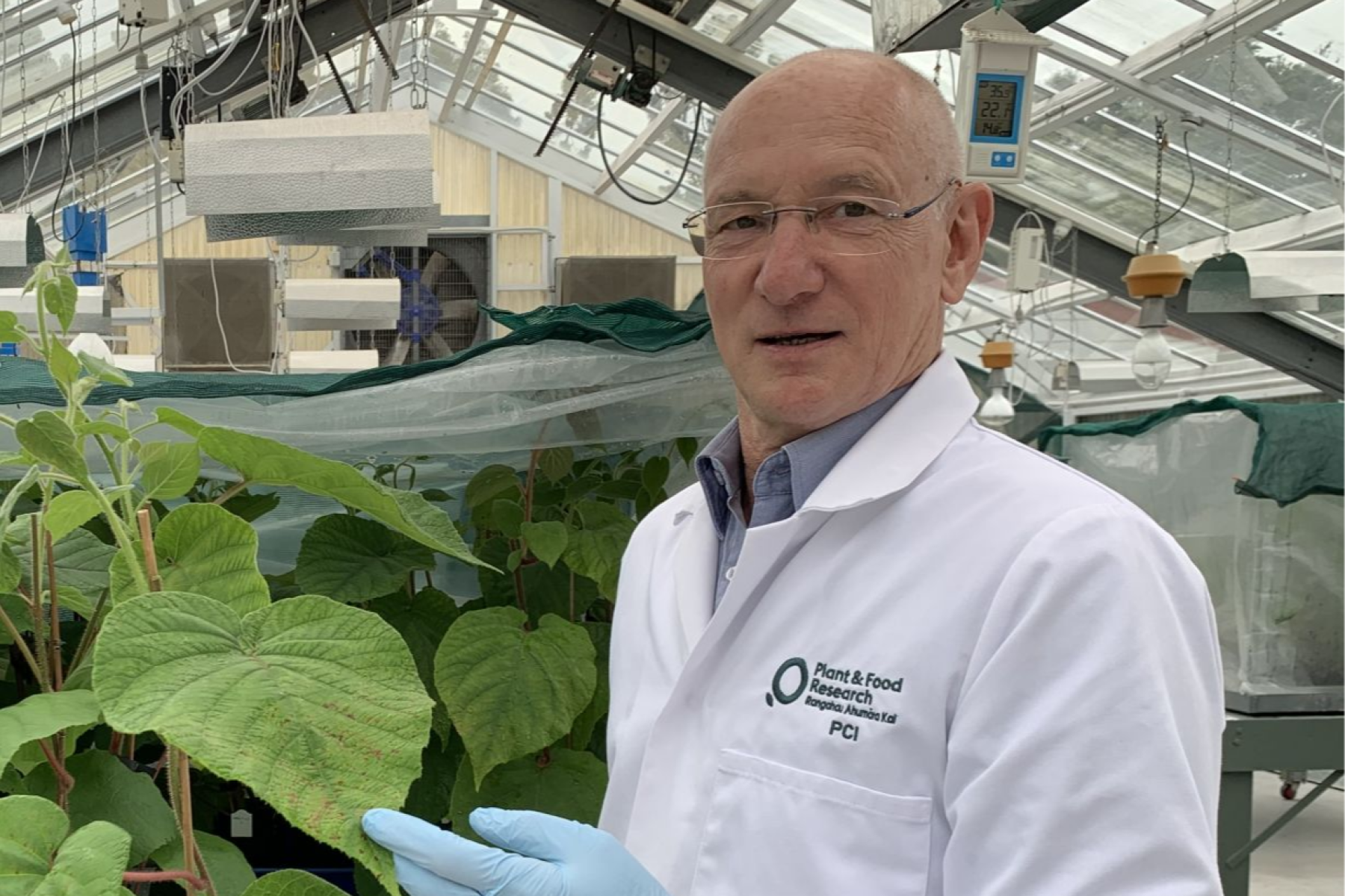 Dr. Joel Vanneste steht in einem Gewächshaus, trägt Arbeitskleidung. Im Hintergrund sind Pflanzen und Laborausrüstung sichtbar.