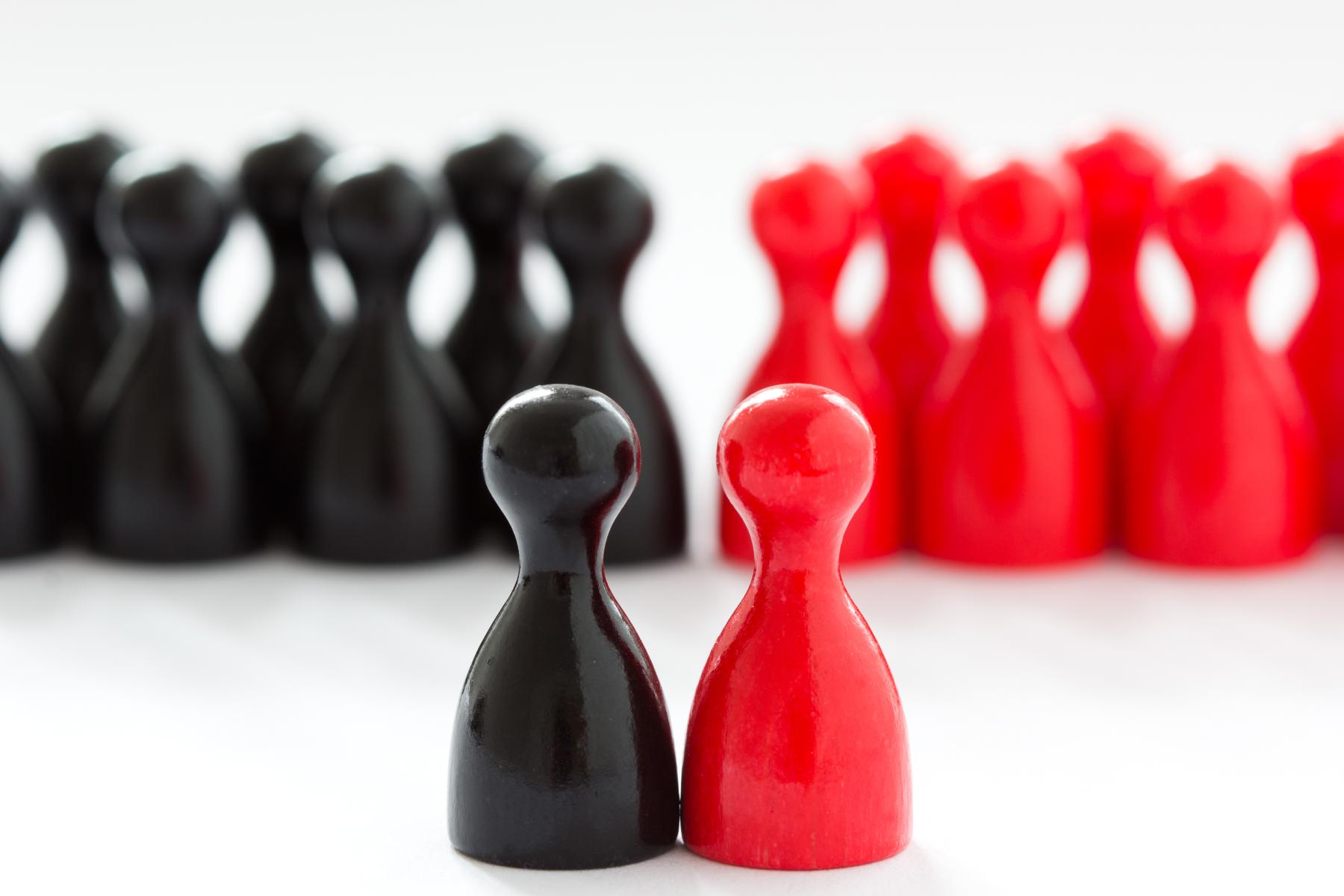 Zwei Spielfiguren, eine schwarz und eine rot, stehen eng beieinander im Vordergrund. Im Hintergrund sind mehrere schwarze und rote Spielfiguren in Gruppen angeordnet.