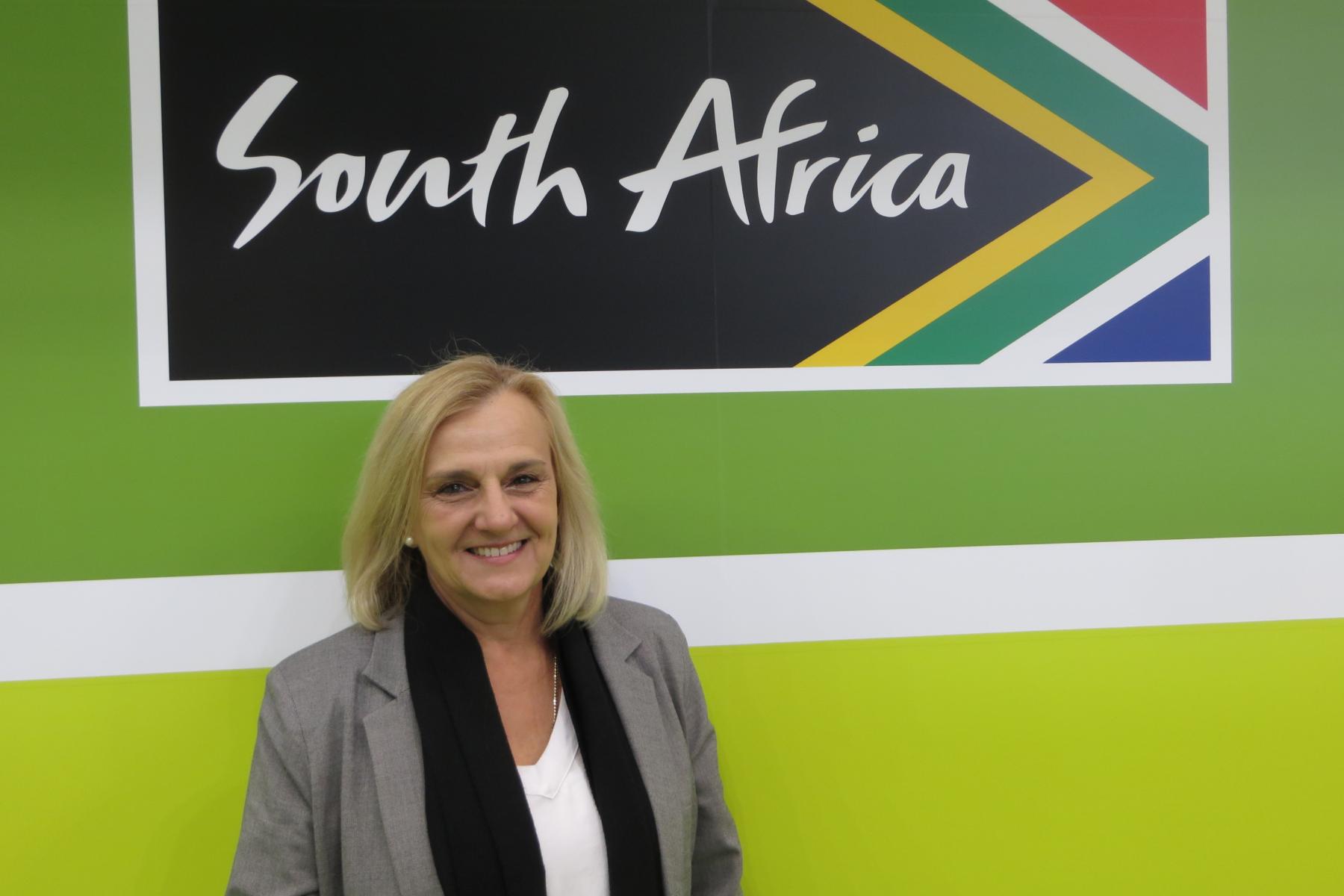 Retha Louw steht vor einer Wand mit dem Schriftzug 'South Africa' und dem südafrikanischen Flaggenmuster in den Farben Grün, Gelb, Blau und Rot.