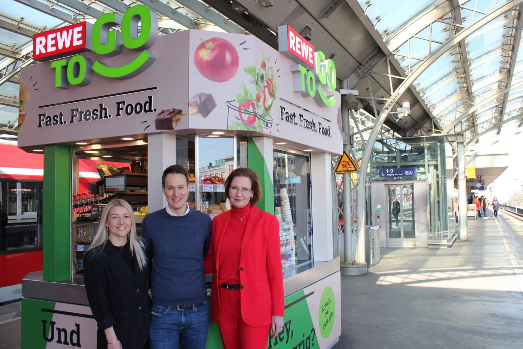 Helen Schnürle, Michael Mayer-Sonnenburg und Jenny Ramme stehen vor einem REWE To Go Kiosk mit Obst- und Lebensmittelangeboten. Der Kiosk ist farbenfroh gestaltet und befindet sich in einem Bahnhof.