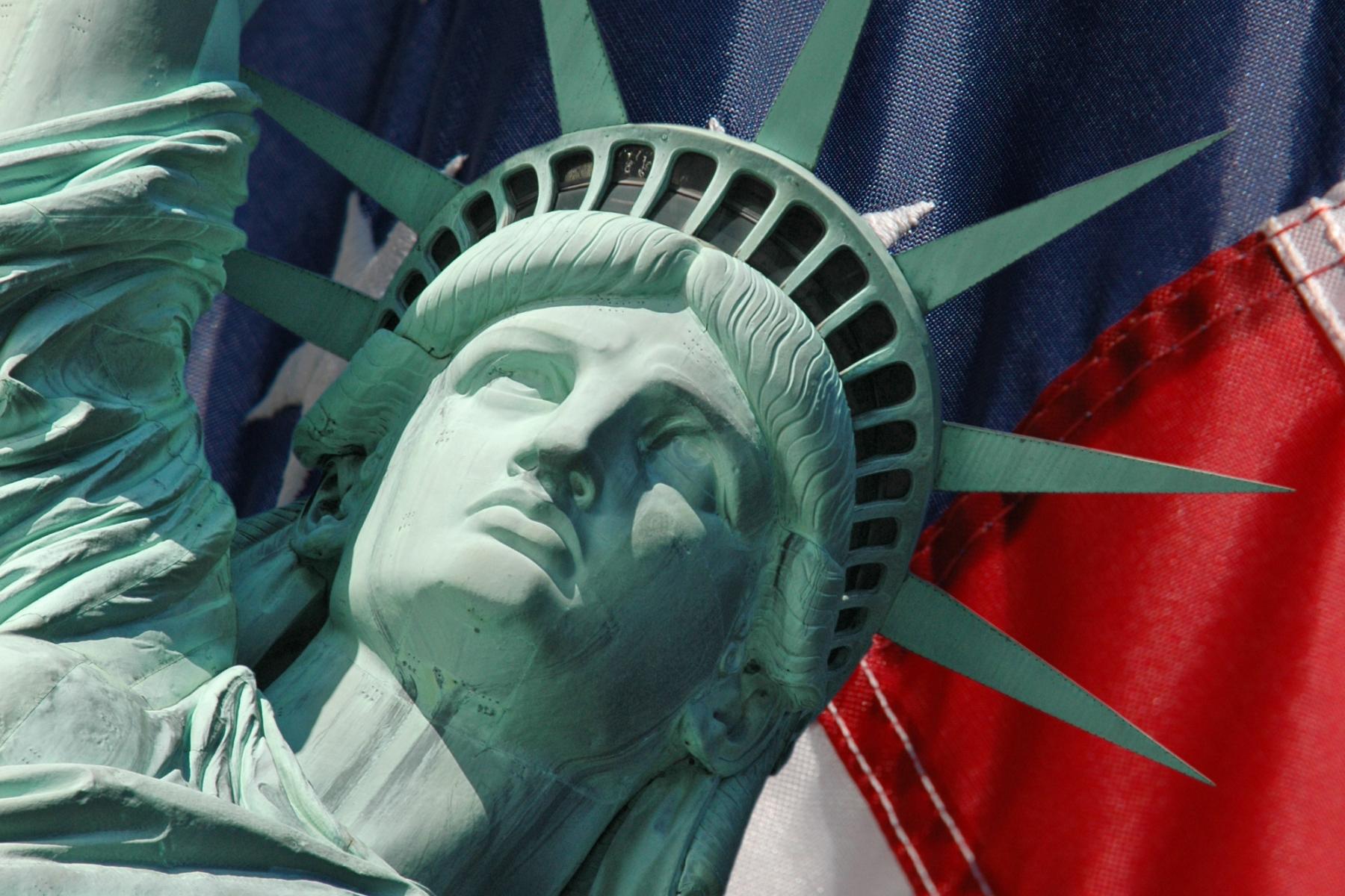 Die Freiheitsstatue mit grünem Kupferfinish, Blick nach oben, vor einem Hintergrund der amerikanischen Flagge mit roten, weißen und blauen Streifen und weißen Sternen.