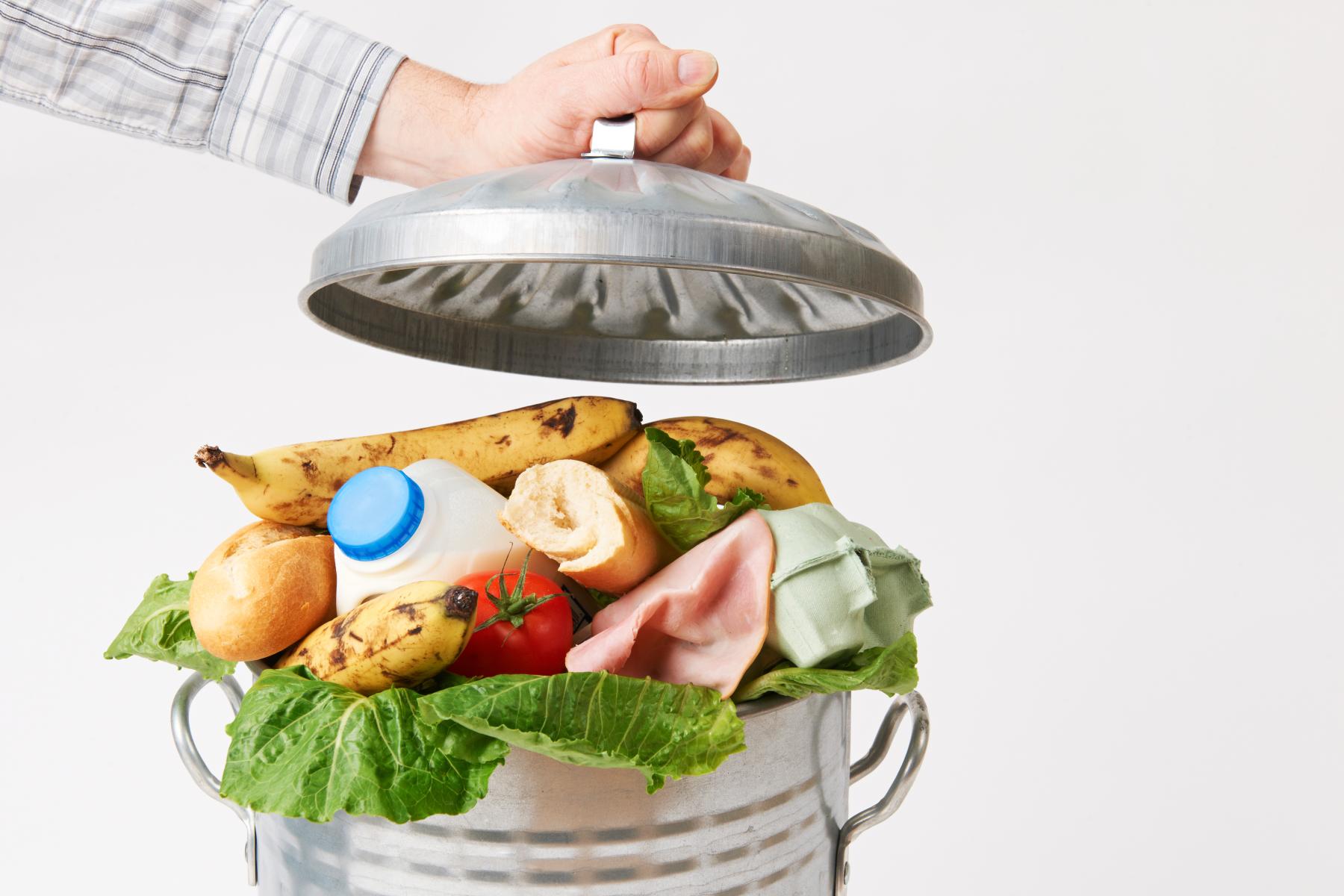 Eine Hand hebt den Deckel eines Metallmüllbehälters, der mit Lebensmitteln gefüllt ist, darunter Bananen, ein Brot, Milch, eine Tomate, Schinken und Salat.