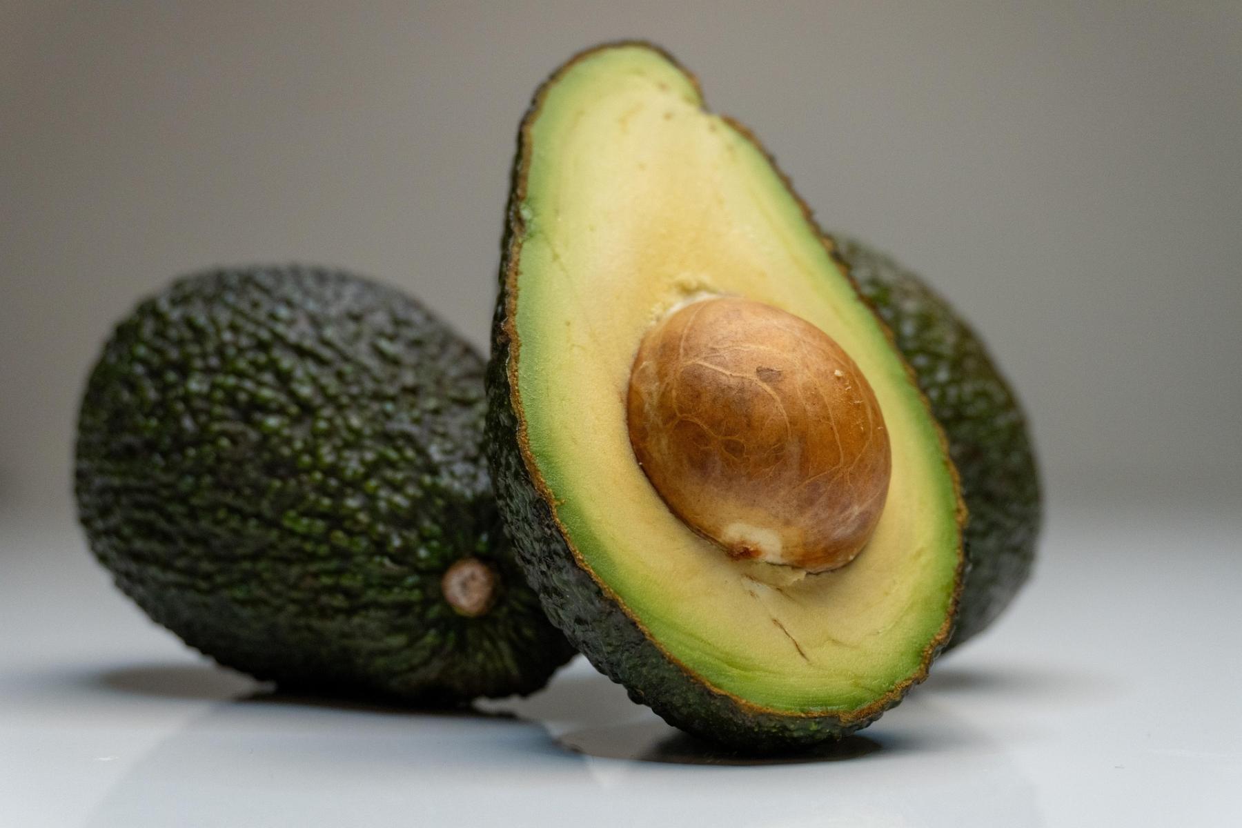 Eine aufgeschnittene Avocado mit einem großen, braunen Kern in der Mitte. Im Hintergrund sind zwei ganze Avocados sichtbar.