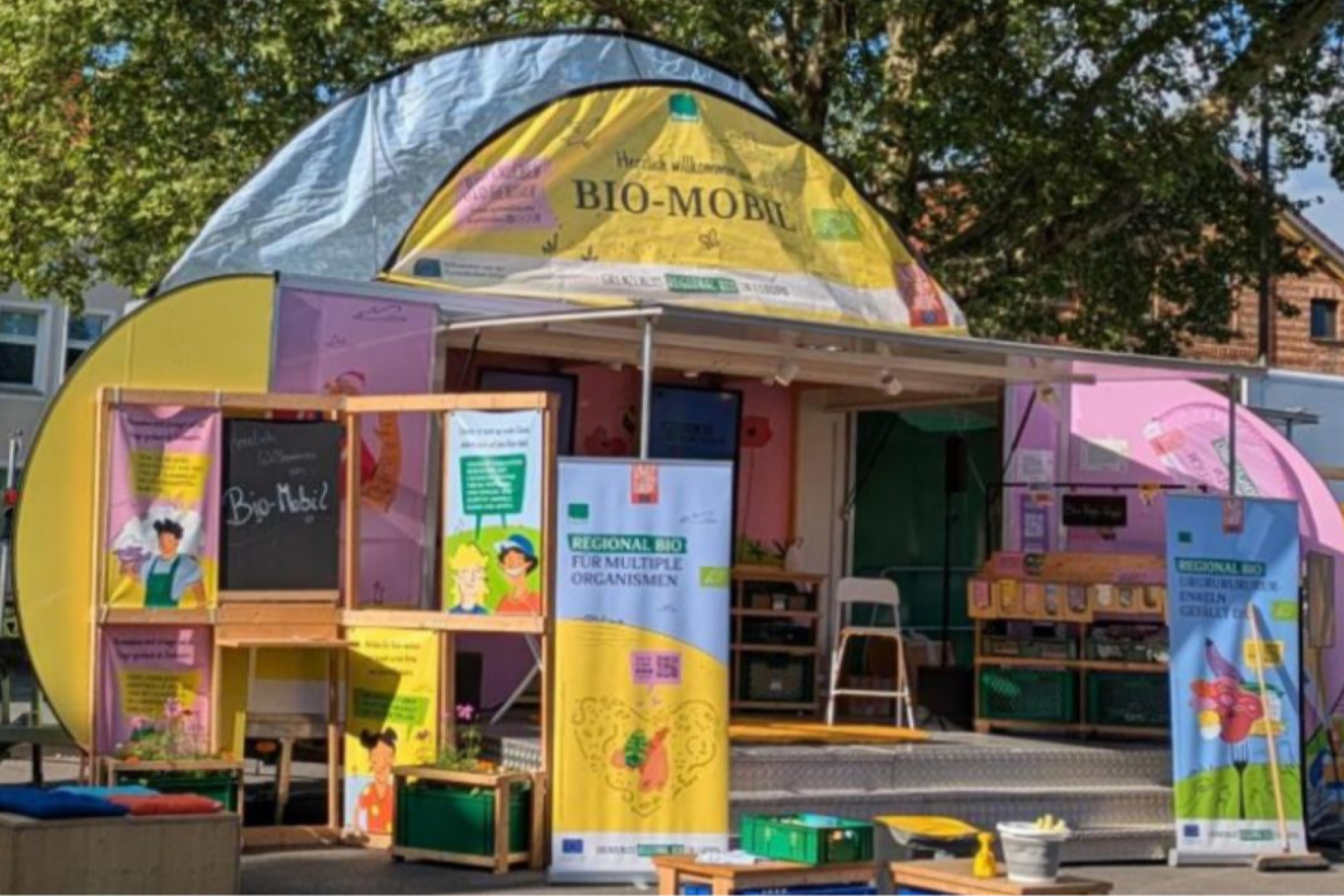 in mobiler Informationsstand mit bunten Wänden in Gelb und Pink, ausgestattet mit Plakaten und einem kleinen Tisch. Auf einer Tafel steht 'Bio-Mobil!'.
