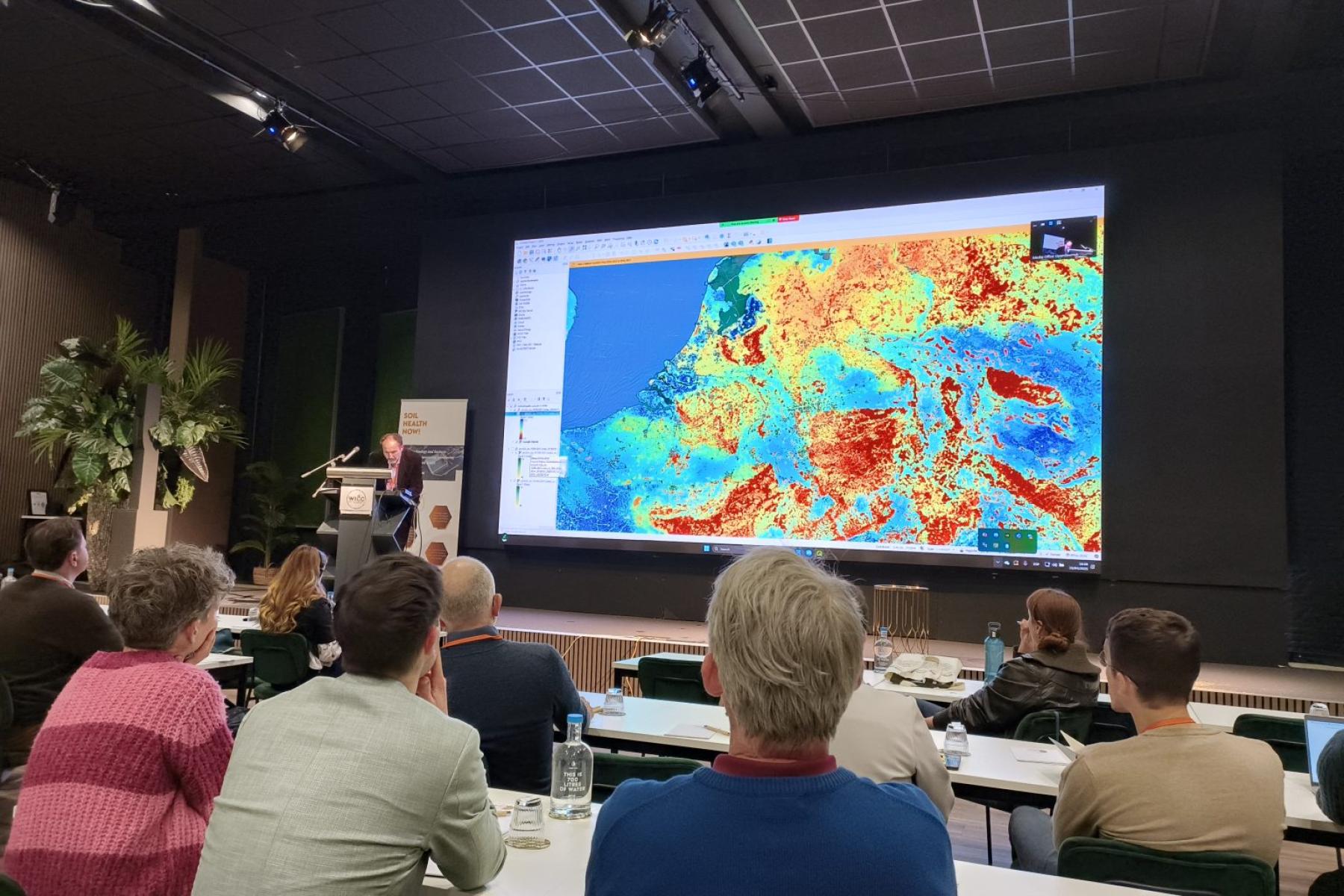 Tomislav Hengl präsentiert farbige Datenvisualisierungen des Soil Health Data Cube auf einer großen Leinwand. Im Vordergrund sind Zuhörer in einem Konferenzraum zu sehen.