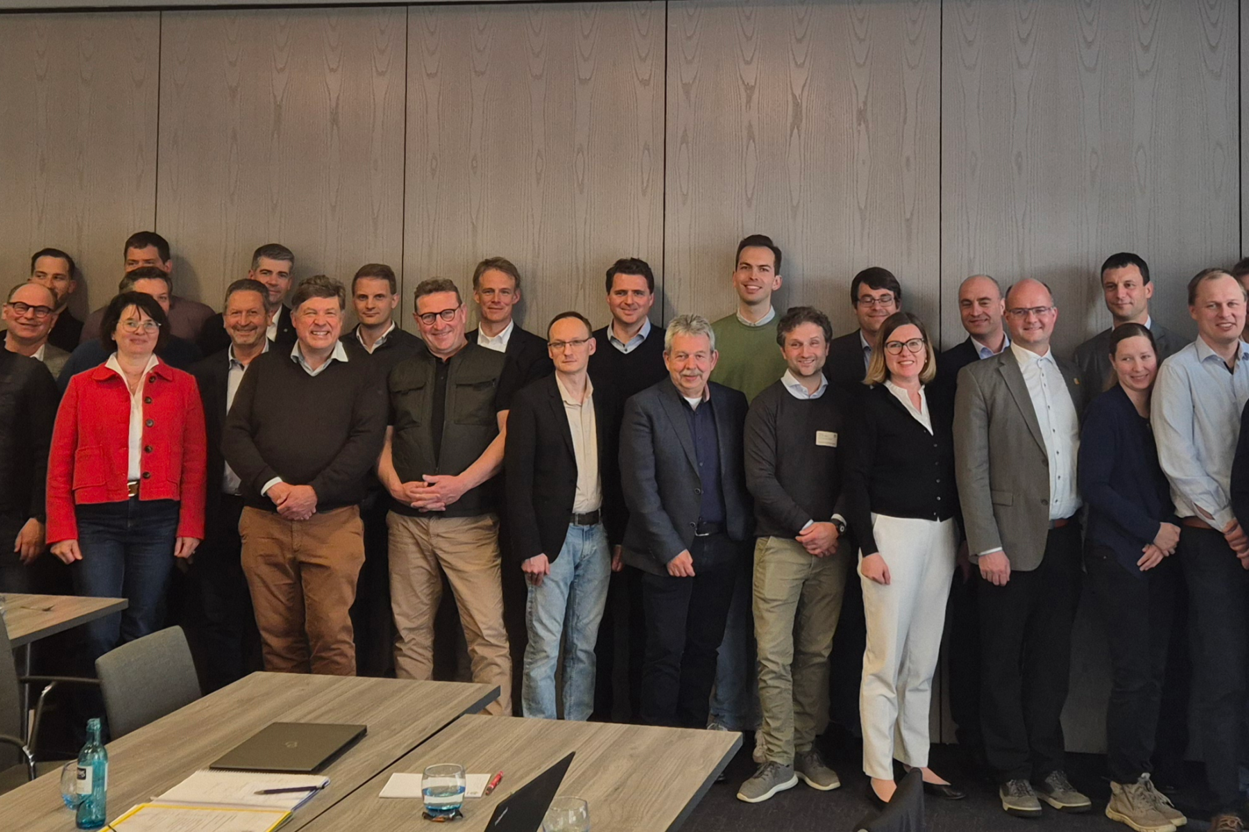 Gruppenfoto von vielen Personen in einem Konferenzraum. Tische mit Unterlagen und Wasserflasche im Vordergrund.