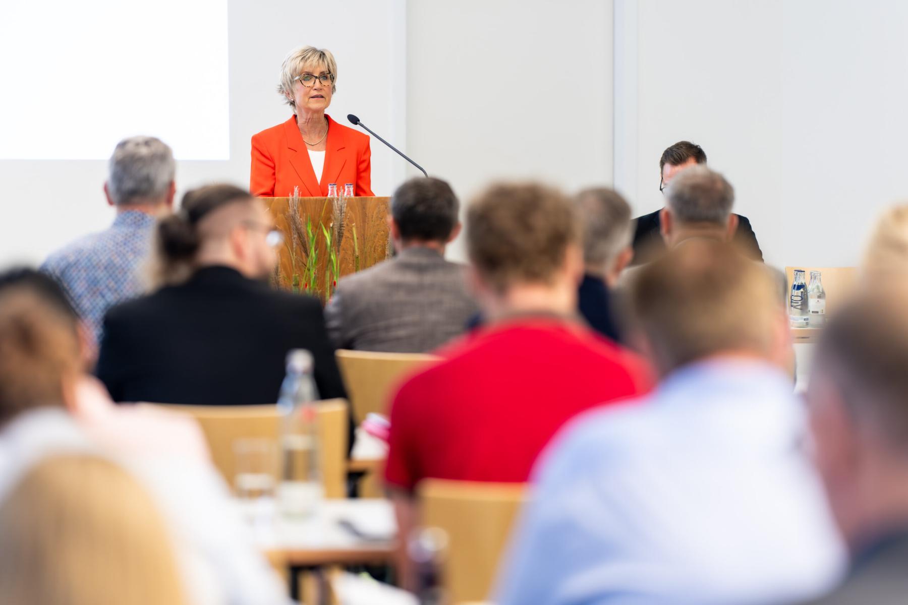 Eva Kähler-Theuerkauf spricht an einem Rednerpult vor einem Publikum. Das Publikum sitzt in Reihen an langen Tischen, auf einem ist auch eine Wasserflasche zu erkennen.