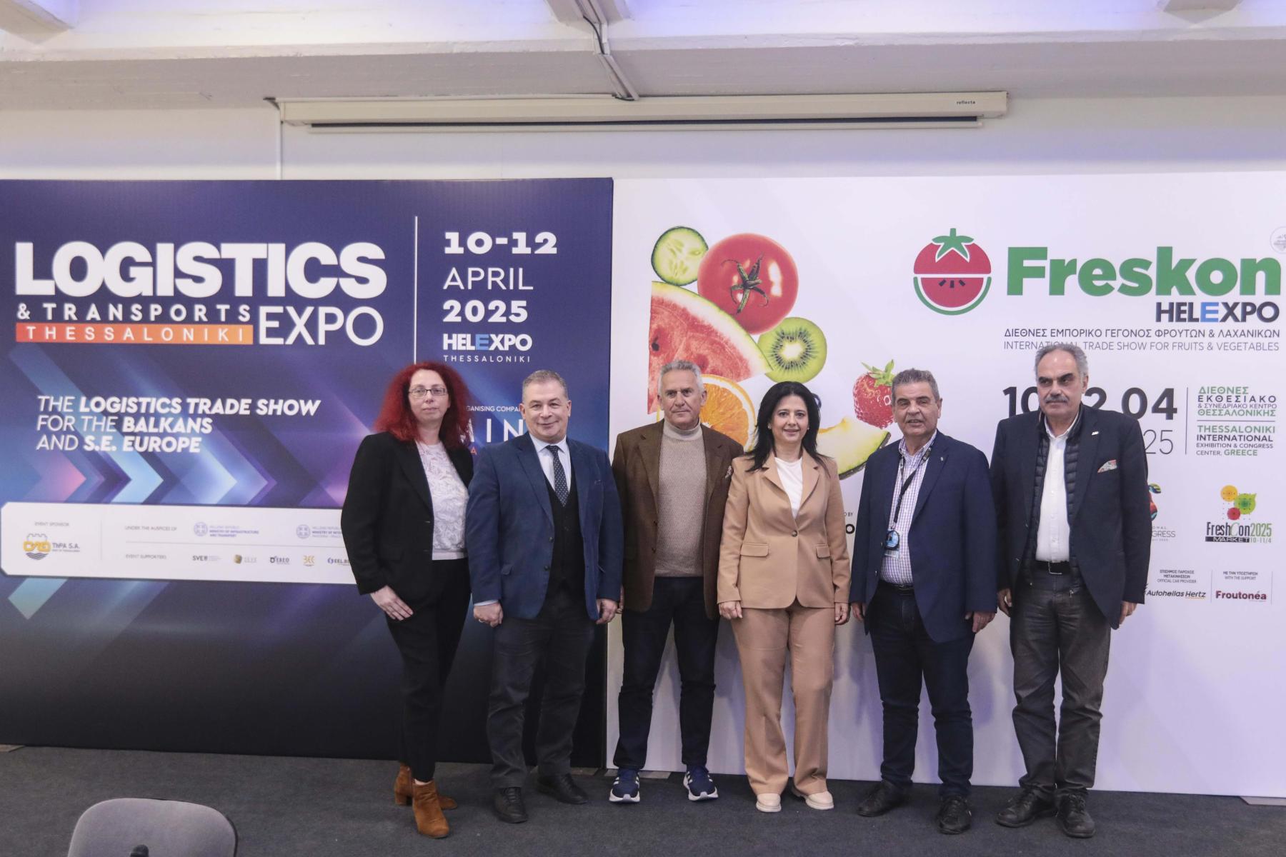 Sechs Personen stehen in einer Reihe vor einer Wand mit zwei Plakaten. Links ein Plakat mit dem Text 'LOGISTICS & TRANSPORTS EXPO'. Rechts ein Plakat mit dem Logo 'Freskon' und dem Datum '10-12 APRIL 2025', sowie Bildern von Obst.