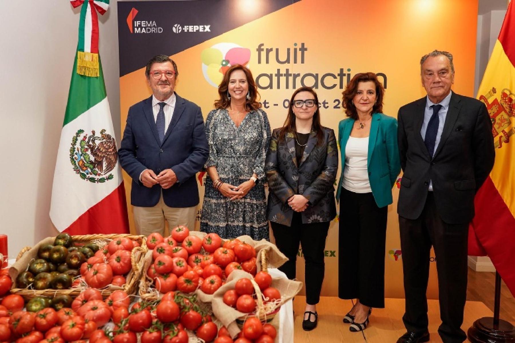 Fünf Personen stehen vor einer Wand mit dem Text 'fruit attraction' und den Flaggen von Mexiko und Spanien. Vor ihnen liegen Körbe mit Tomaten auf einer Präsentationsfläche
