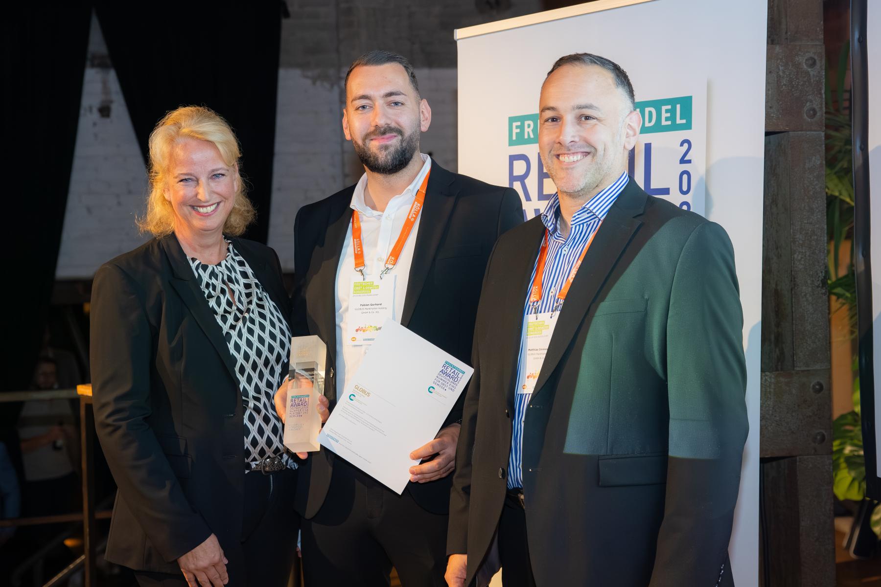 Drei Personen in Businesskleidung bei der Verleihung des Retail Award 2025. Ein Vertreter der GLOBUS Markthallen Holding GmbH & Co. KG hält eine Urkunde und einen Award. Im Hintergrund ist ein Roll-Up mit der Aufschrift „Retail Award“ sichtbar.