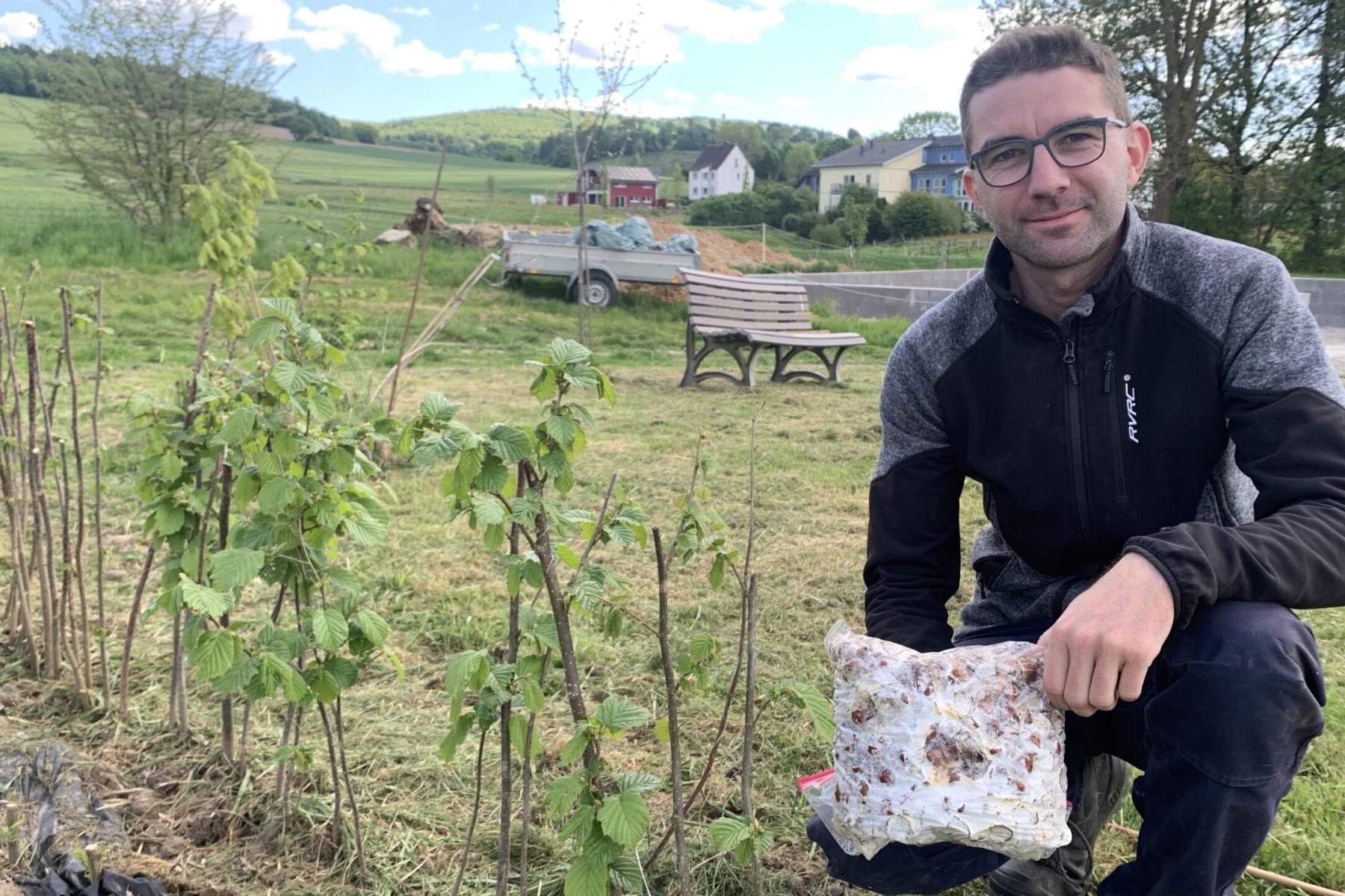 Hendrik Hoffmann kniet im Freien und hält ein Paket mit Pflanzenmaterial in der Hand. Im Hintergrund sind Sträucher und ein Wohnhaus sichtbar.