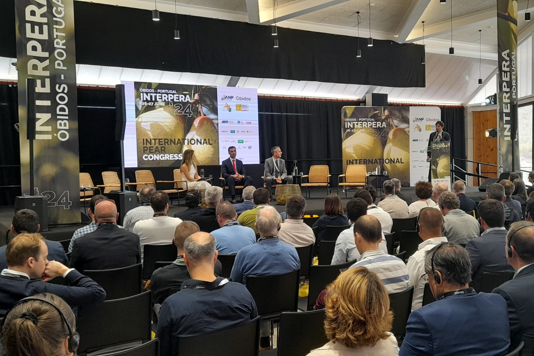 Blick auf eine Bühne mit mehreren Personen im Publikum. Auf der Bühne sitzen drei Personen und eine weitere steht rechts am Rand und spricht am Mikrofon. Im Hintergrund ein Banner mit 'INTERPERA 24 INTERNATIONAL PEAR CONGRESS'.