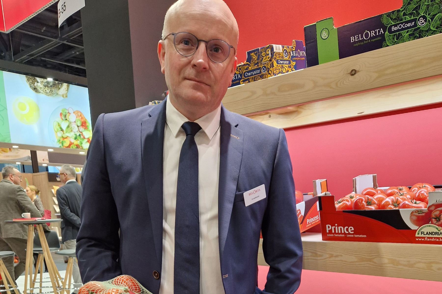 Jo Lambrecht hält ein Netz mit Tomaten, steht vor einem Regal mit weiteren Tomatenkartons bei einem Messestand.