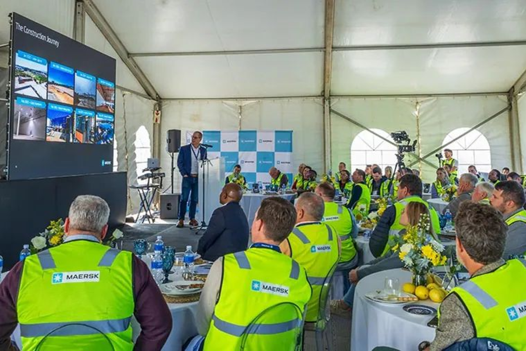 Eine Präsentation in einem Zelt mit einem Redner vor einer Leinwand. Die Zuhörer tragen gelbe Maersk-Sicherheitswesten. 