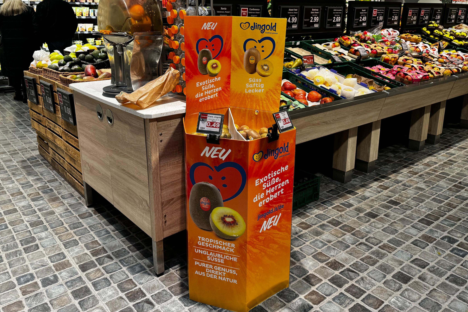 Ein Verkaufsstand von Jingold mit einer Auswahl an Kiwis. Der Stand zeigt die Aufschrift 'NEU' und Informationen zu tropischen Früchten. Im Hintergrund sind Obst- und Gemüseregale sichtbar.