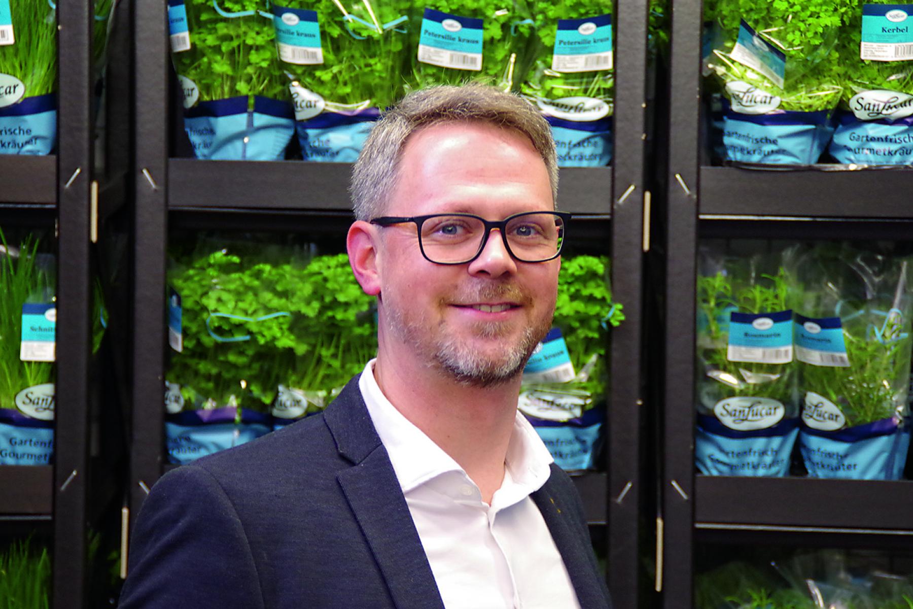 Nils Doerwald steht vor einem Regal mit frischen Kräutern in Töpfen, die in blauen Verpackungen von 'Sangrar' präsentiert werden.