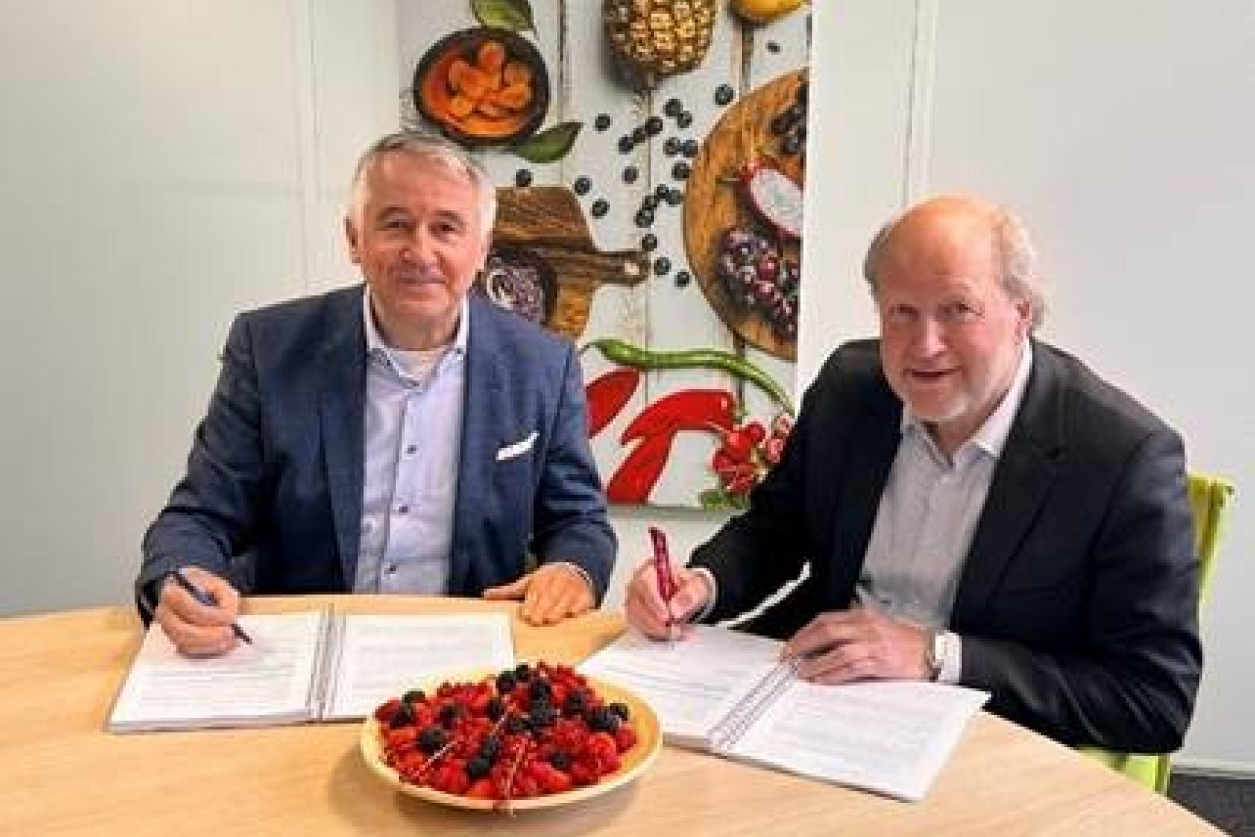 Richard Schouten und René van der Burg sitzen an einem Tisch und unterschreiben Dokumente. Vor ihnen steht eine Schüssel mit Beeren.