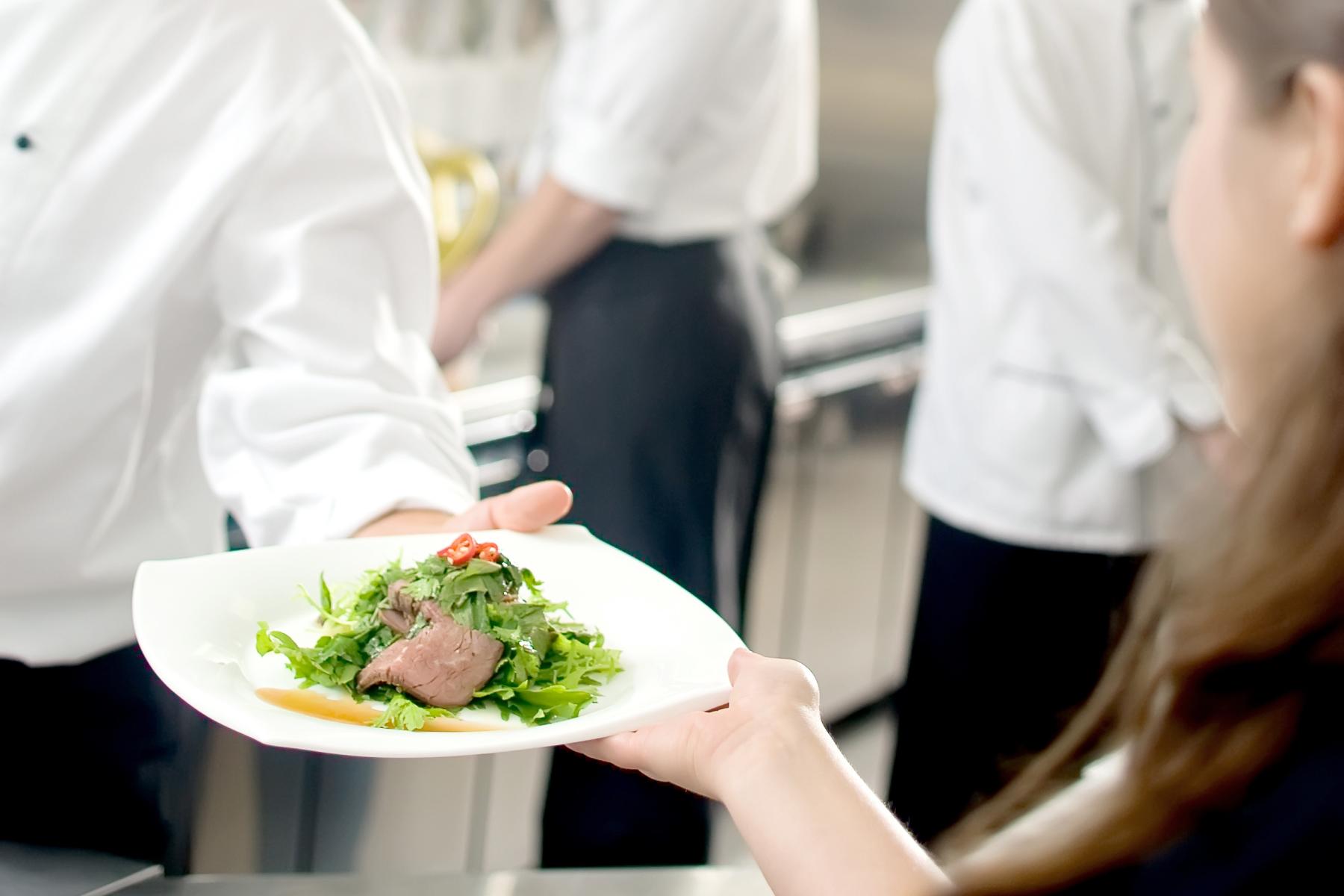 Ein Koch in einer weißen Kochjacke reicht einen Teller mit Fleisch und Salat an eine Person. Im Hintergrund sind weitere Köche zu sehen.