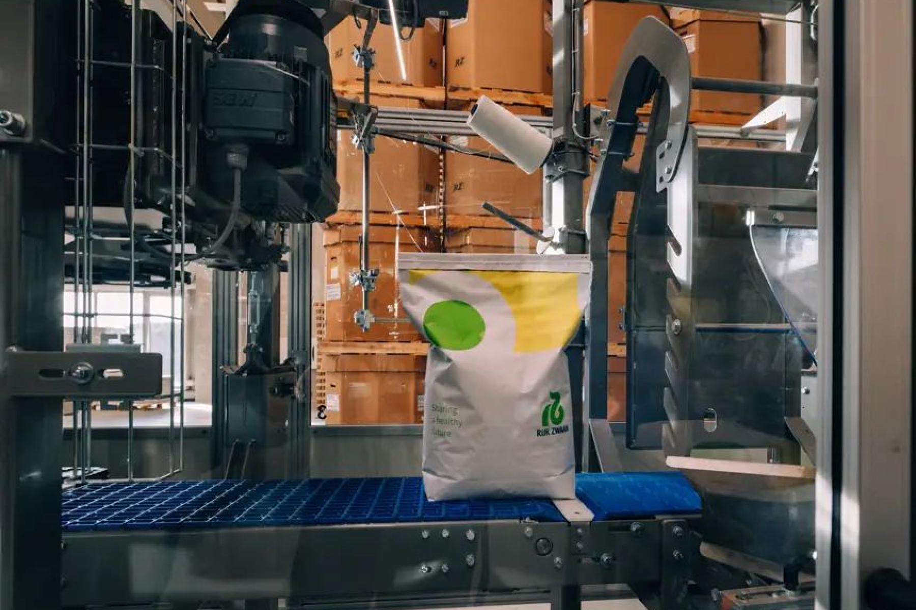 Eine Maschine verpackt einen weißen Sack mit grünem und gelbem Logo von Rijk Zwaan. Im Hintergrund sind braune Kartons sichtbar.