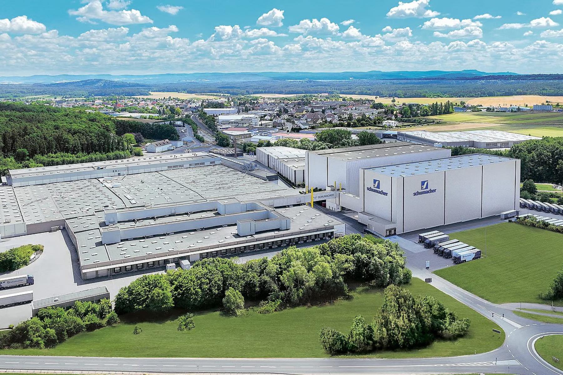 Luftaufnahme der Schumacher Packaging-Werke mit mehreren Dächern, umgeben von Wäldern und Feldern. Im Vordergrund sind Lkw und eine Straße sichtbar.