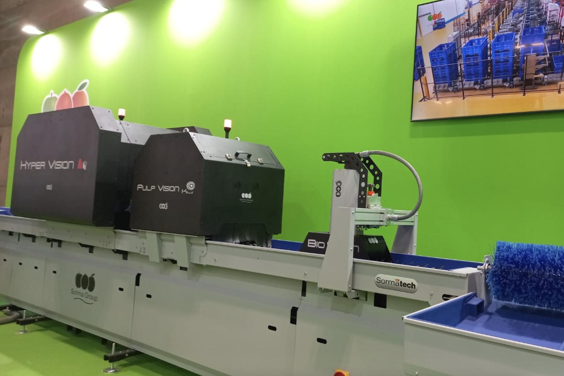Maschine zur Fruchtsortierung mit zwei großen schwarzen Geräten, beschriftet mit 'HYPER VISION' und 'PULP VISION', auf einem Förderband. Im Hintergrund ein Bildschirm mit blauen Kisten.