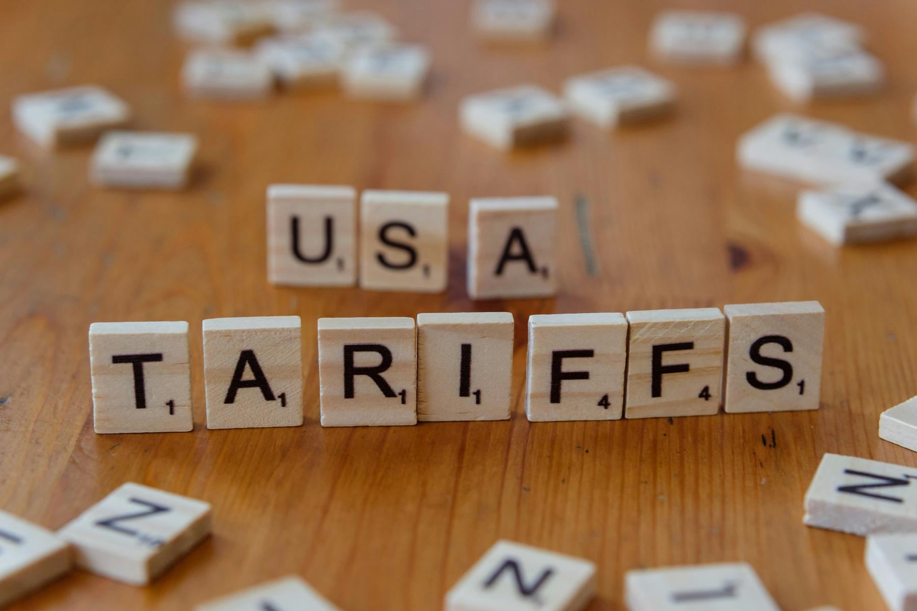 Holzbuchstaben auf einem Tisch bilden das Wort 'TARIFFS' und 'USA'. Die Buchstaben sind in einem Scrabble-ähnlichen Stil angeordnet.