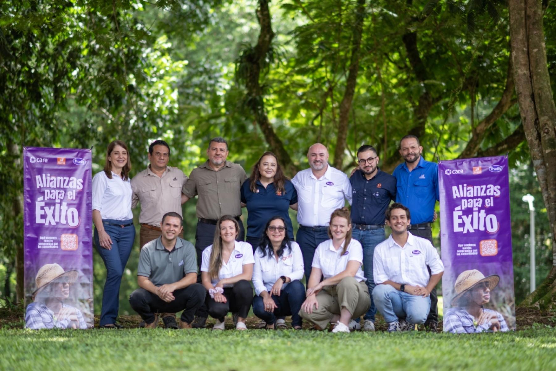Gruppenfoto von 12 Personen im Freien, umgeben von Bäumen. Im Hintergrund zwei Banner mit dem Text 'Alianzas para el Éxito'.