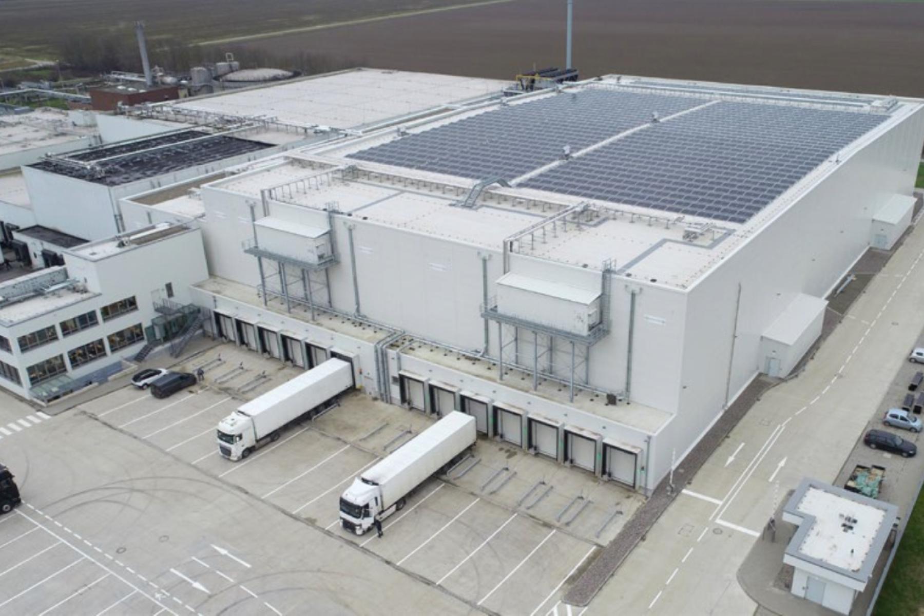 Luftaufnahme eines großen Lagergebäudes mit Solarpanelen auf dem Dach. Vor dem Gebäude stehen mehrere Lkw an den Ladebuchten.