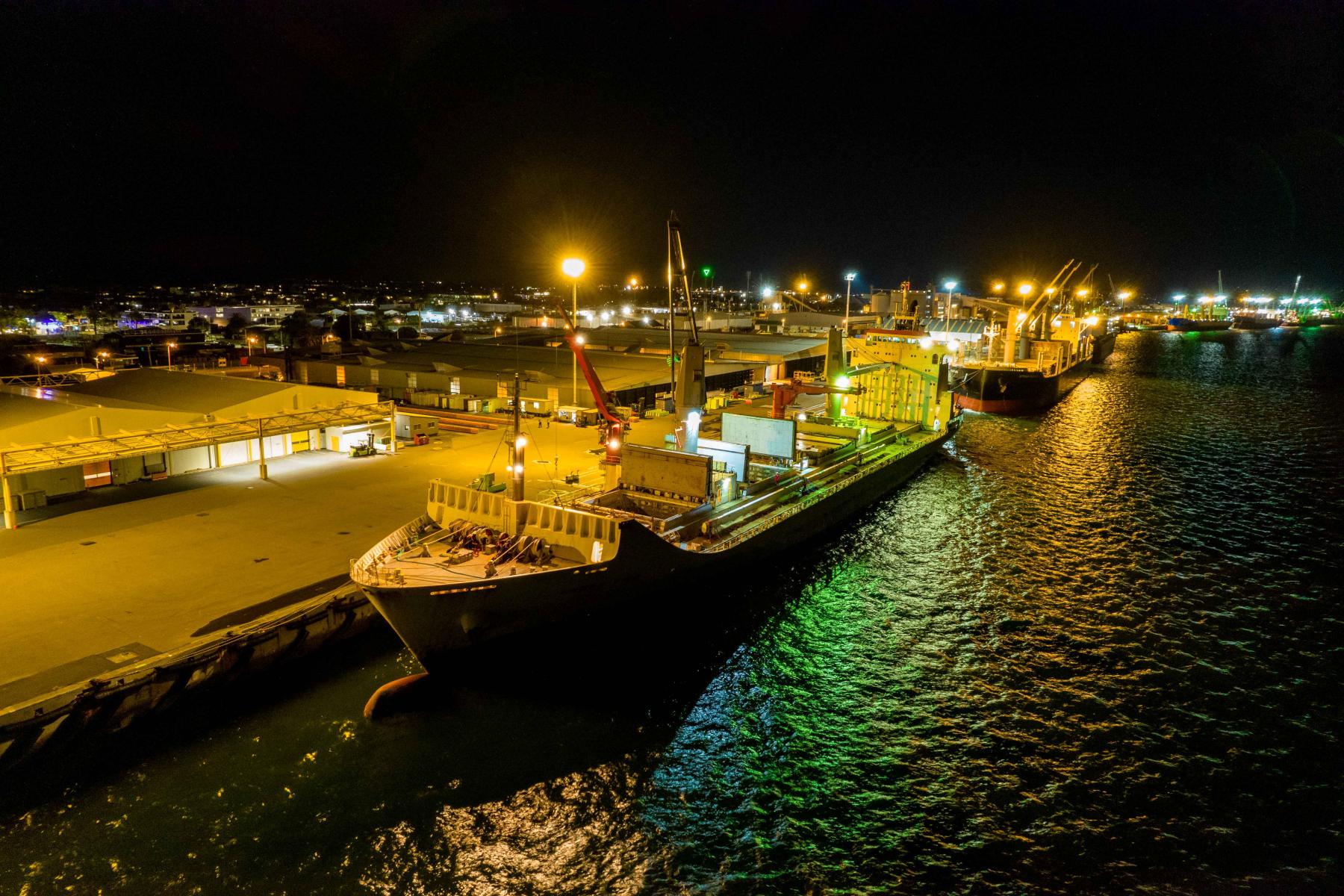Großes Frachtschiff mit geöffneten Frachtklappen im Hafen bei Nacht. Beleuchtete Hafenanlagen im Hintergrund.