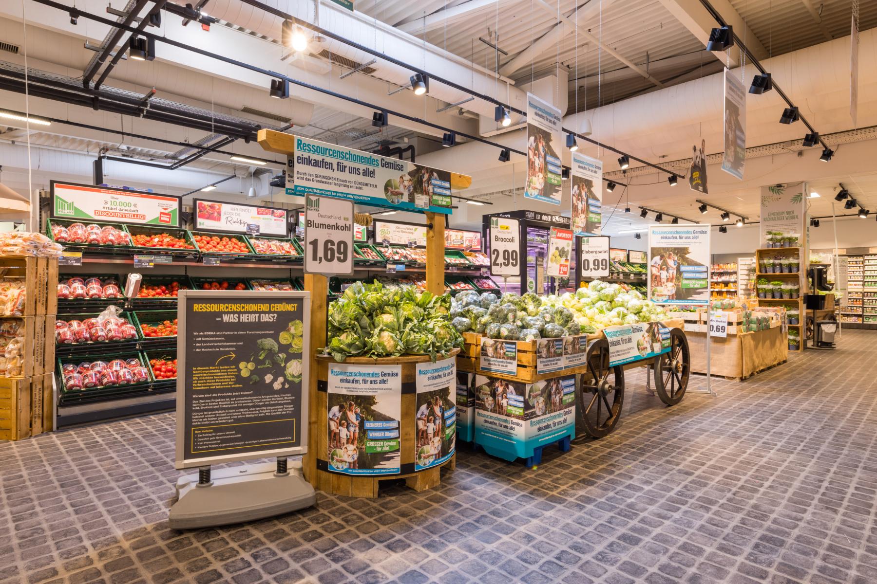 Verkaufsfläche mit Regalen und Wagen voller Blumenkohl, Preisschilder und Informationsplakate in einem Supermarkt.