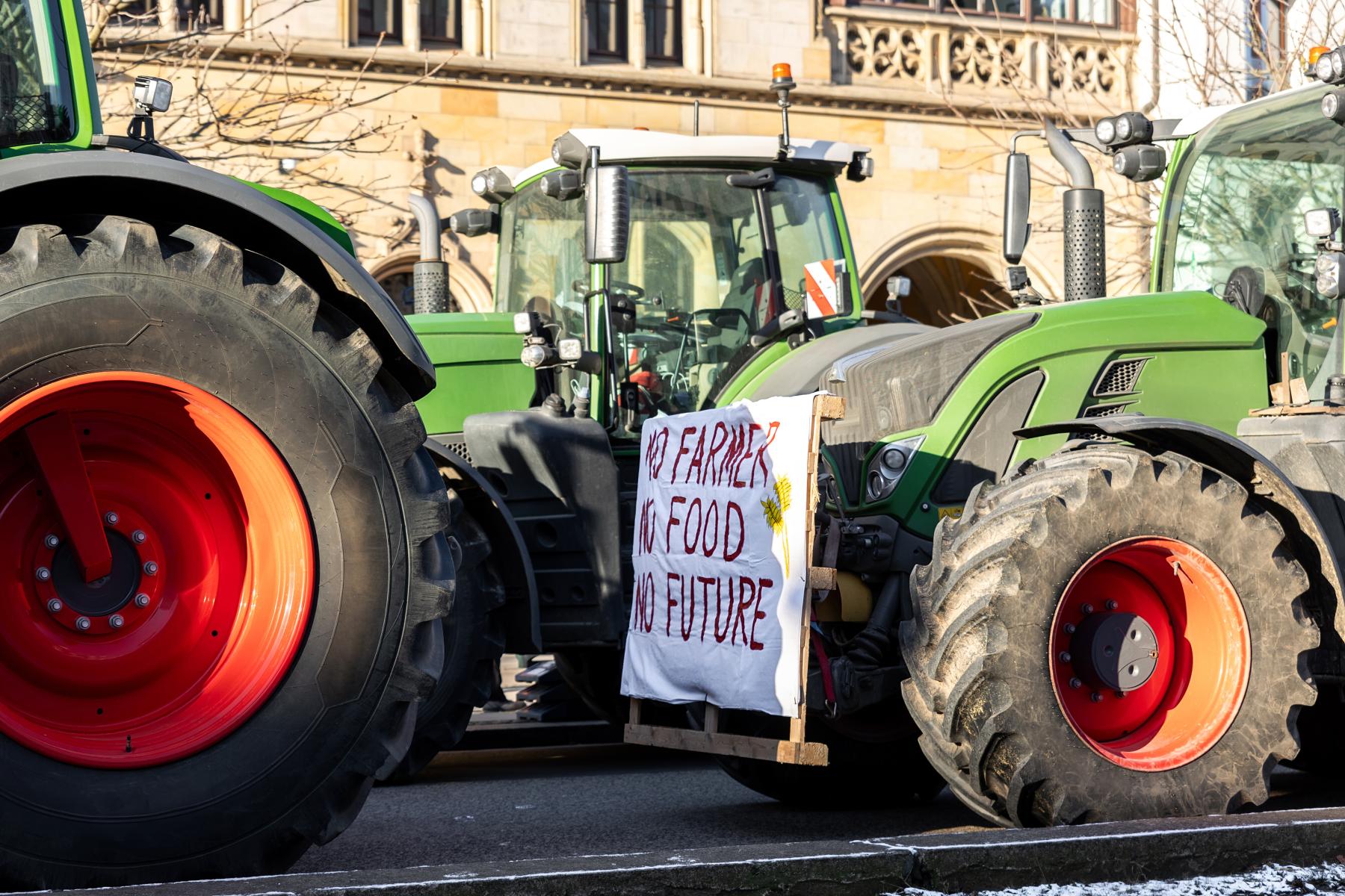 Grüne Traktoren auf Straße, an einem ein weißes Transparent mit rotem Text 'NO FARMER NO FOOD NO FUTURE'
