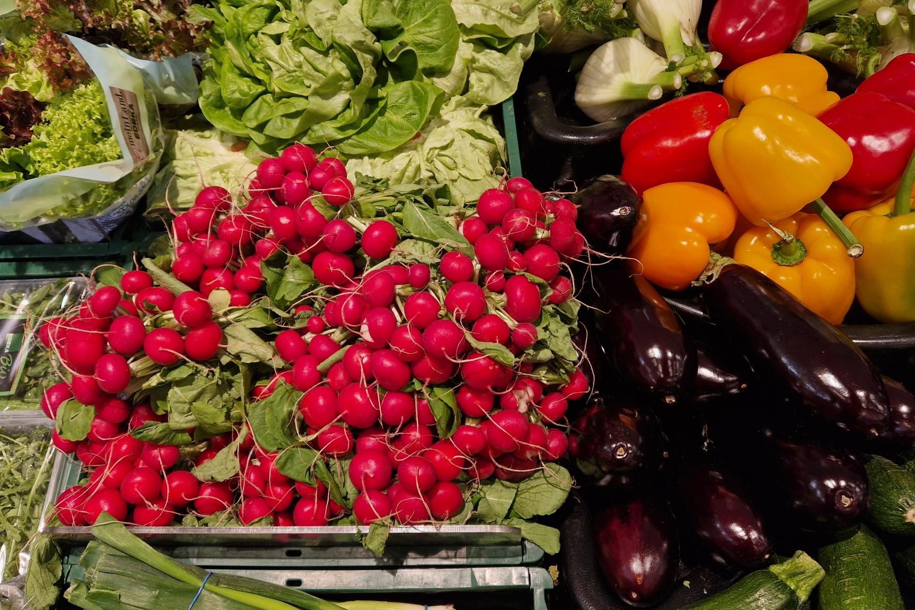 Frische, rote Radieschen mit grünen Blättern, daneben bunte Paprika in Gelb, Orange und Rot, Auberginen und Salatköpfe in einer Obst- und Gemüseabteilung.