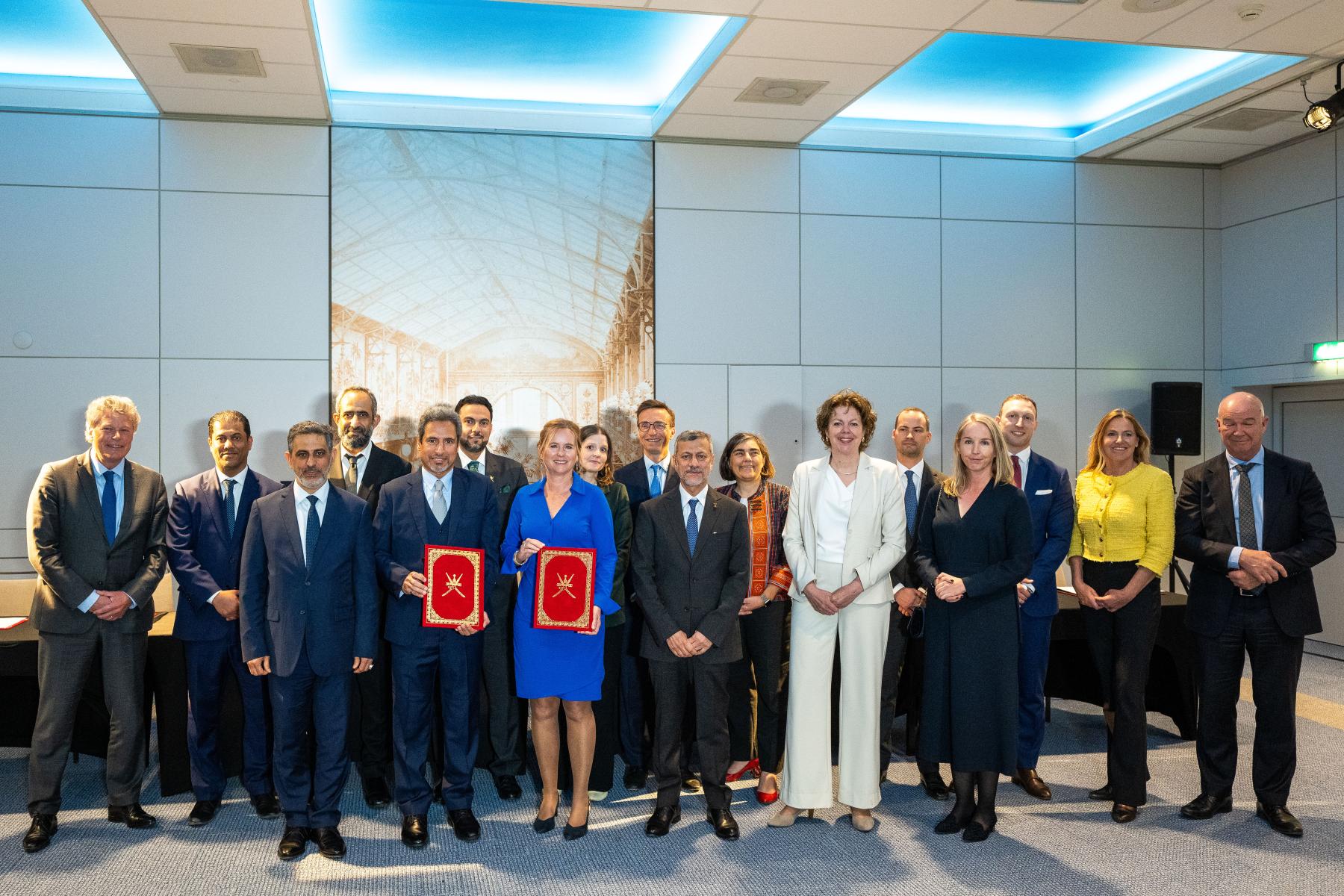 Gruppe von Personen in formeller Kleidung, zwei halten rote Ordner mit goldenen Emblemen, Hintergrund mit großem Bild einer Halle und blauer Deckenbeleuchtung.