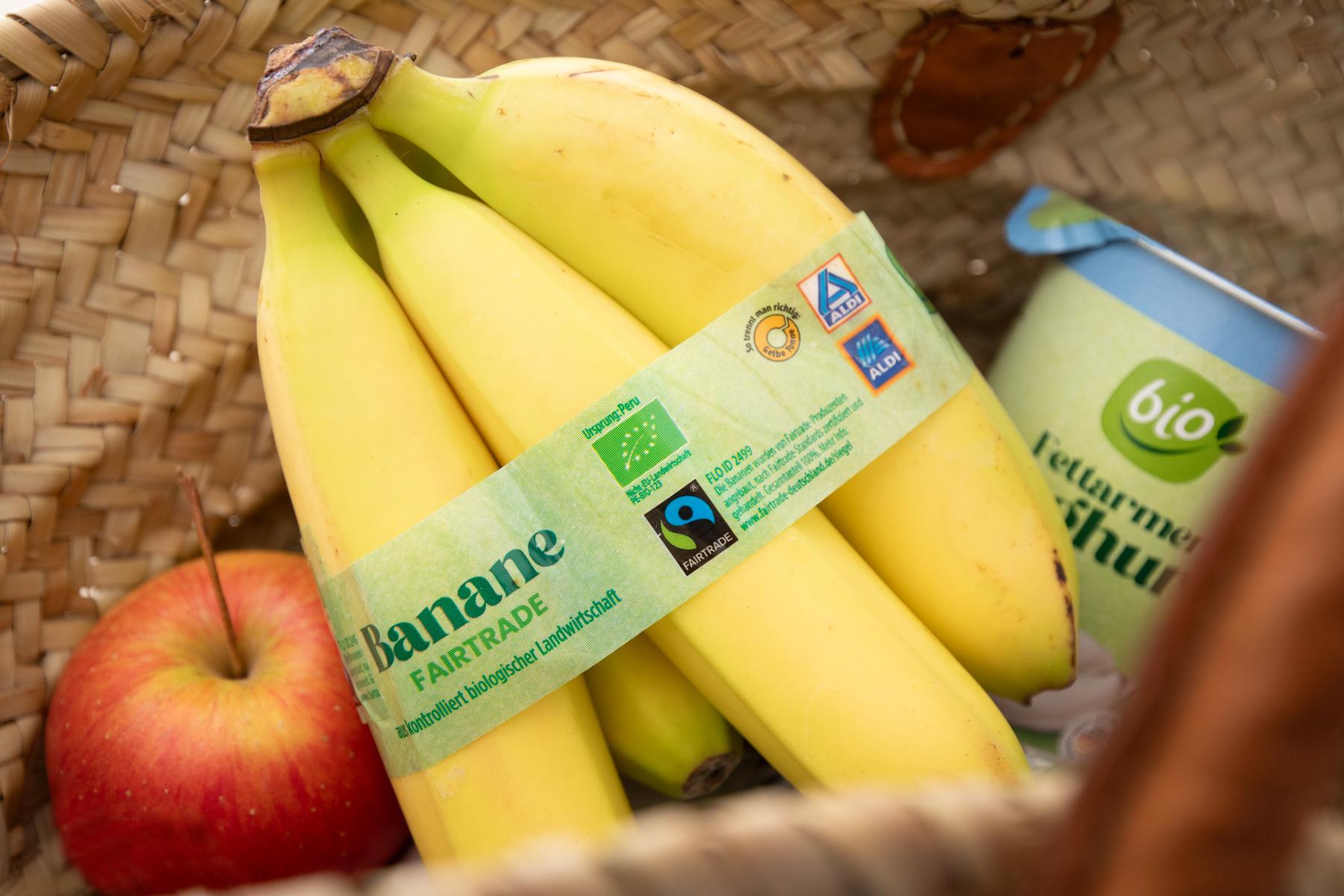 Bündel gelber Bananen mit Fairtrade-Etikett, daneben roter Apfel und Becher mit grünem Bio-Logo in geflochtenem Korb.