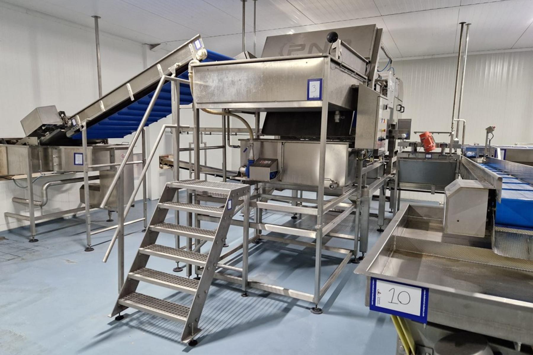 Große Edelstahlmaschine mit Treppe, Förderbändern und mehreren nummerierten Stationen in einer Produktionshalle.