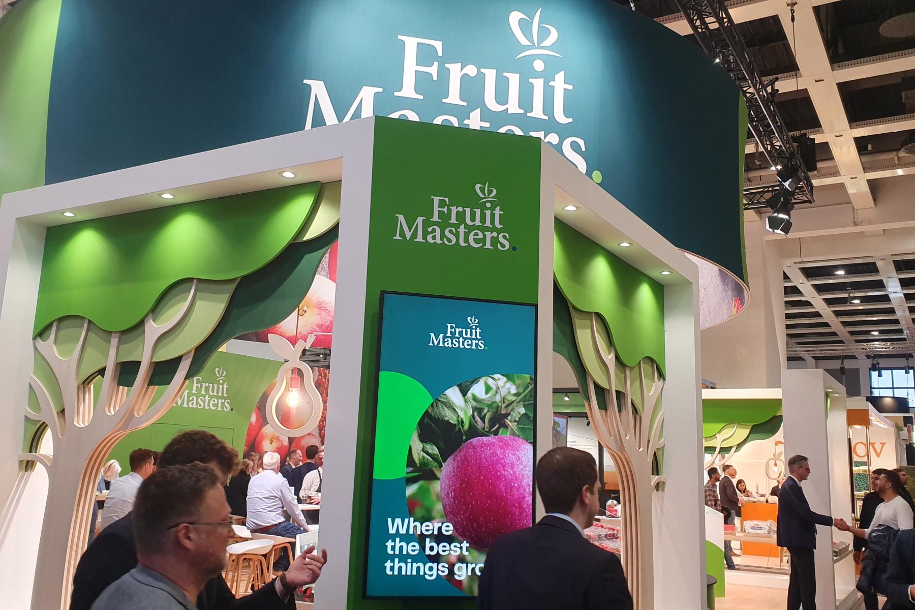 Messestand von Fruit Masters mit grün-weißer Gestaltung, Baum-Dekorationen und digitalem Bildschirm mit Text 'Where the best things grow'.