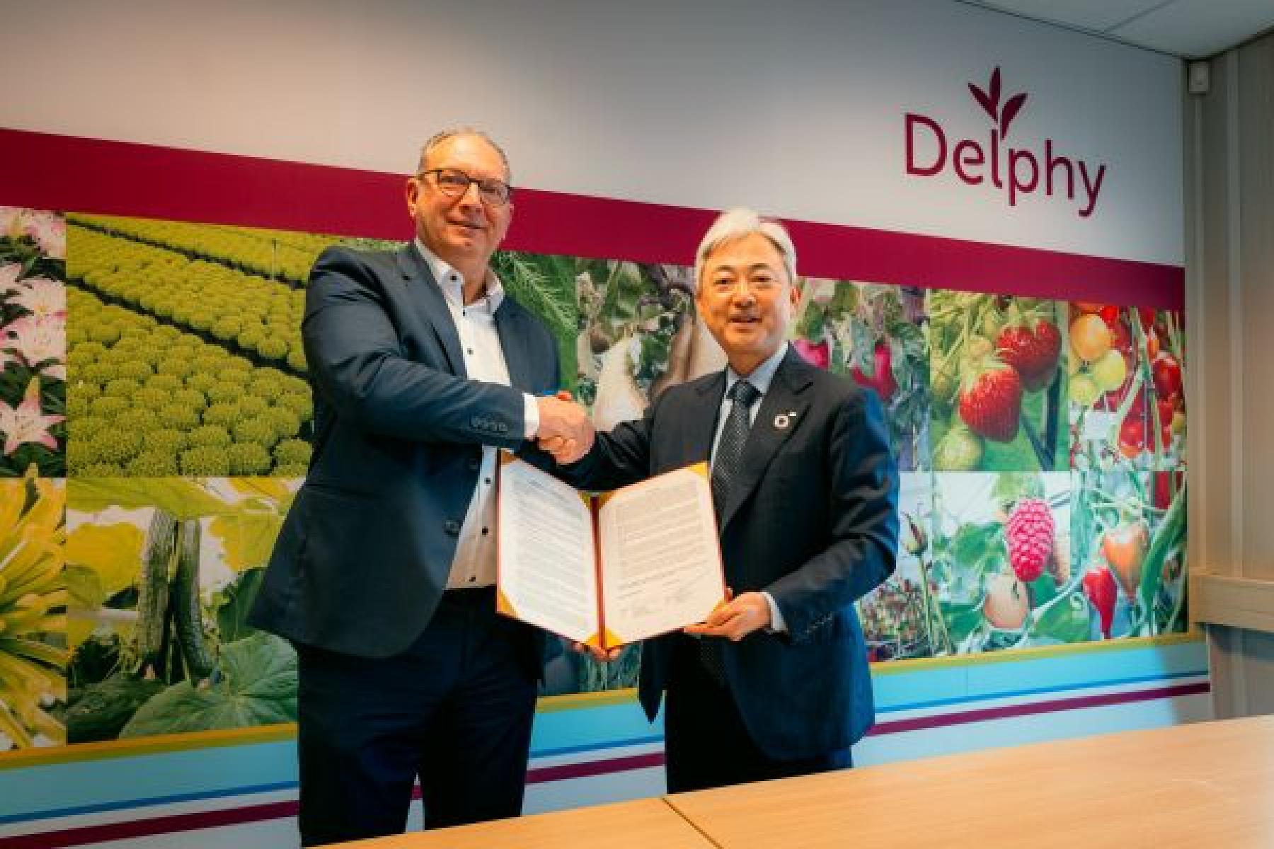 Zwei Männer in Anzügen schütteln sich die Hand, einer hält ein geöffnetes Dokument, im Hintergrund bunte Bilder von Obst und Gemüse und das Logo 'Delphy'.