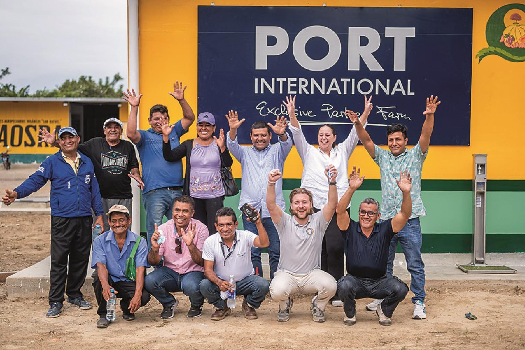 Gruppe von Menschen vor gelb-grünem Gebäude mit großem Schild 'PORT INTERNATIONAL Exclusive Partner Farm', einige mit erhobenen Händen.