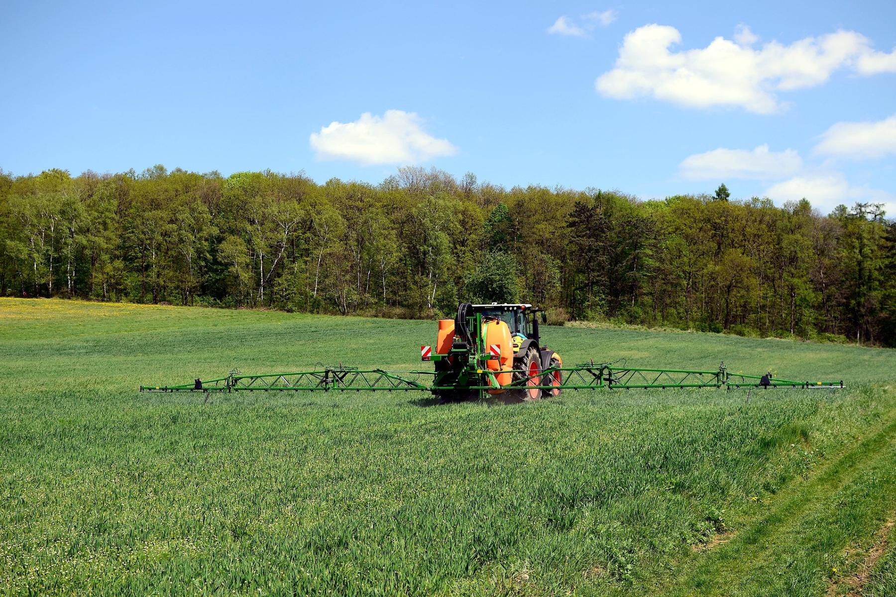 Traktor mit breitem Pflanzenschutzgerät fährt über grünes Feld vor Wald und blauem Himmel mit Wolken.