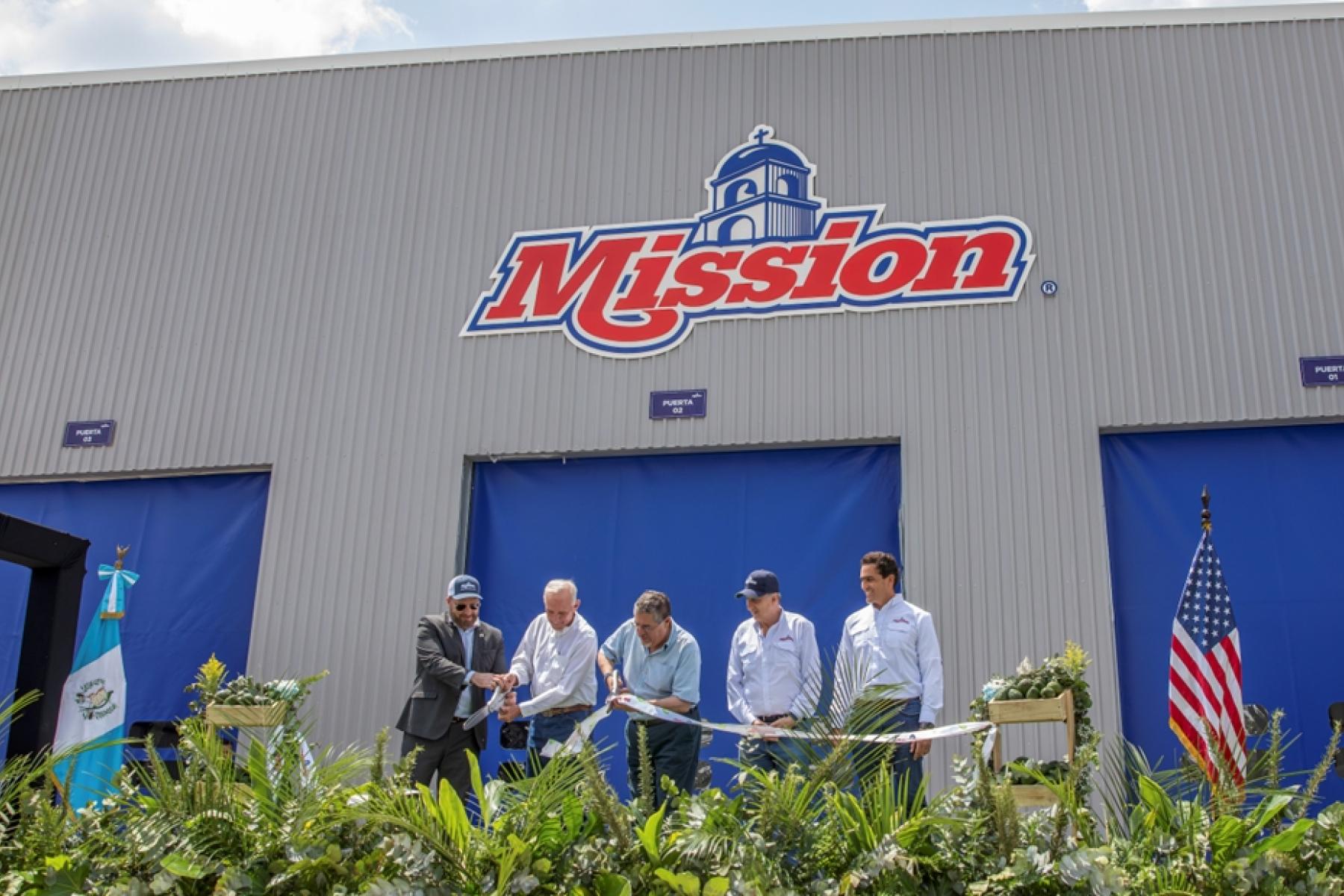 Fünf Männer stehen vor einem Gebäude mit dem Schriftzug 'Mission' und halten ein Band, umgeben von Pflanzen, guatemaltekischer und US-amerikanischer Flagge.